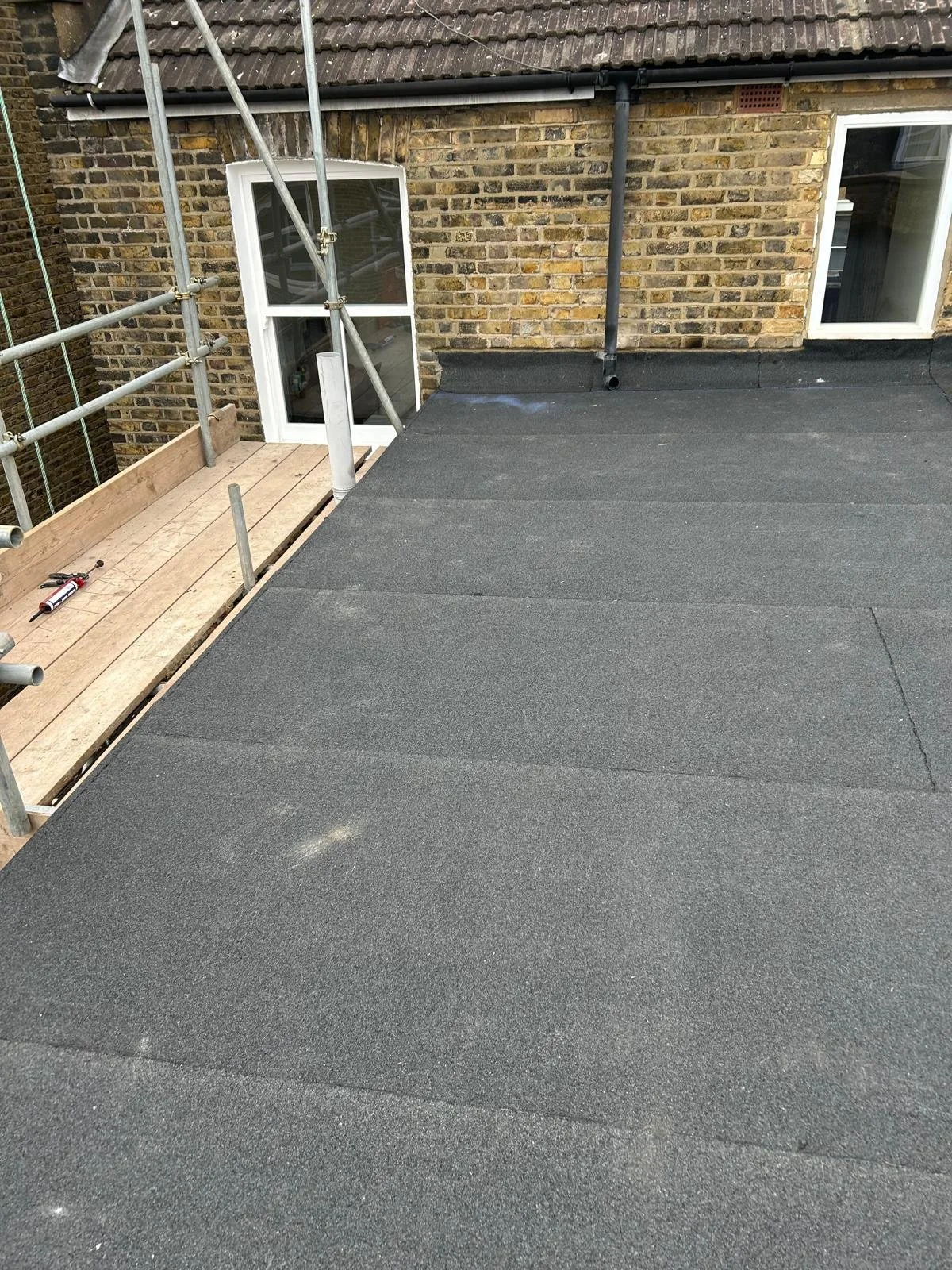 A flat rooftop with black roofing material next to a brick house wall with white-framed windows and scaffolding under construction.