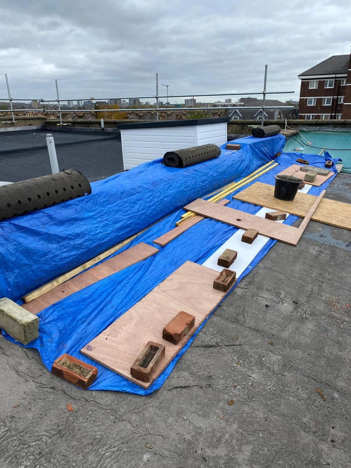 Construction materials on a rooftop, including blue tarp, wooden boards, bricks, black foam rollers, and tools, with an overcast sky and city skyline in the background.