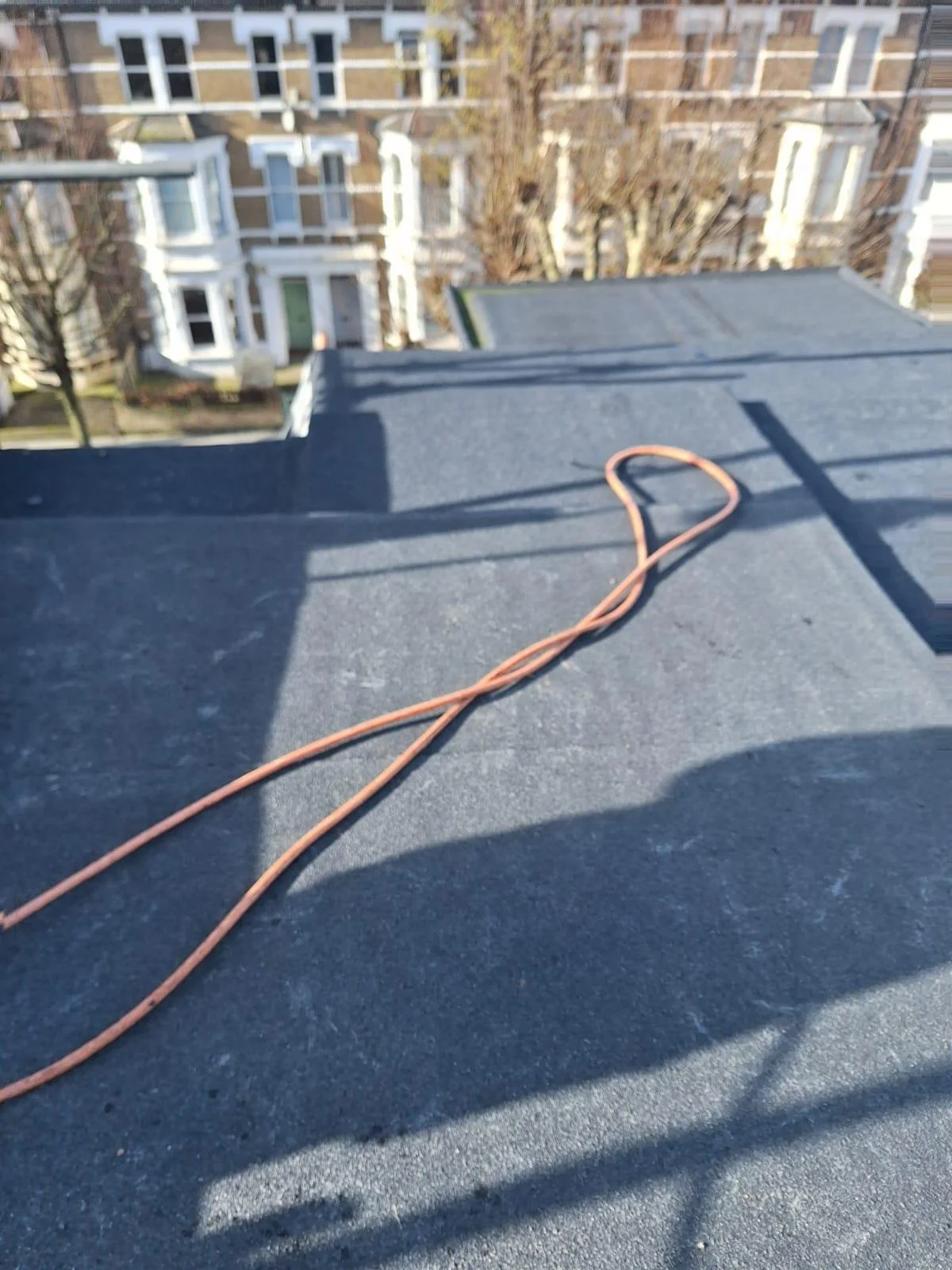 A rooftop with a black surface and an orange extension cord lying on it, with neighboring rooftops, trees, and residential buildings in the background.