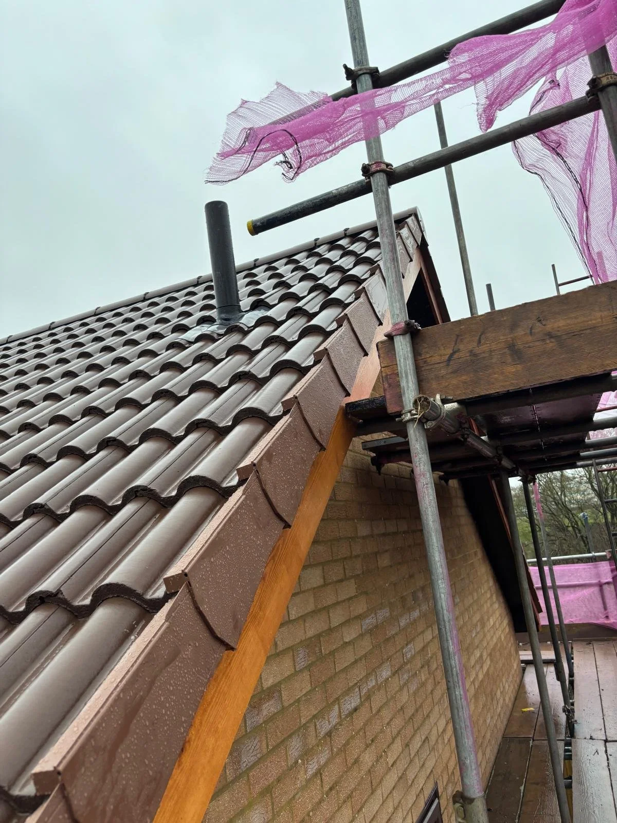 Construction scaffolding on a building with a tiled roof, showing part of the roof edge, chimney pipe, and scaffolding with pink netting.
