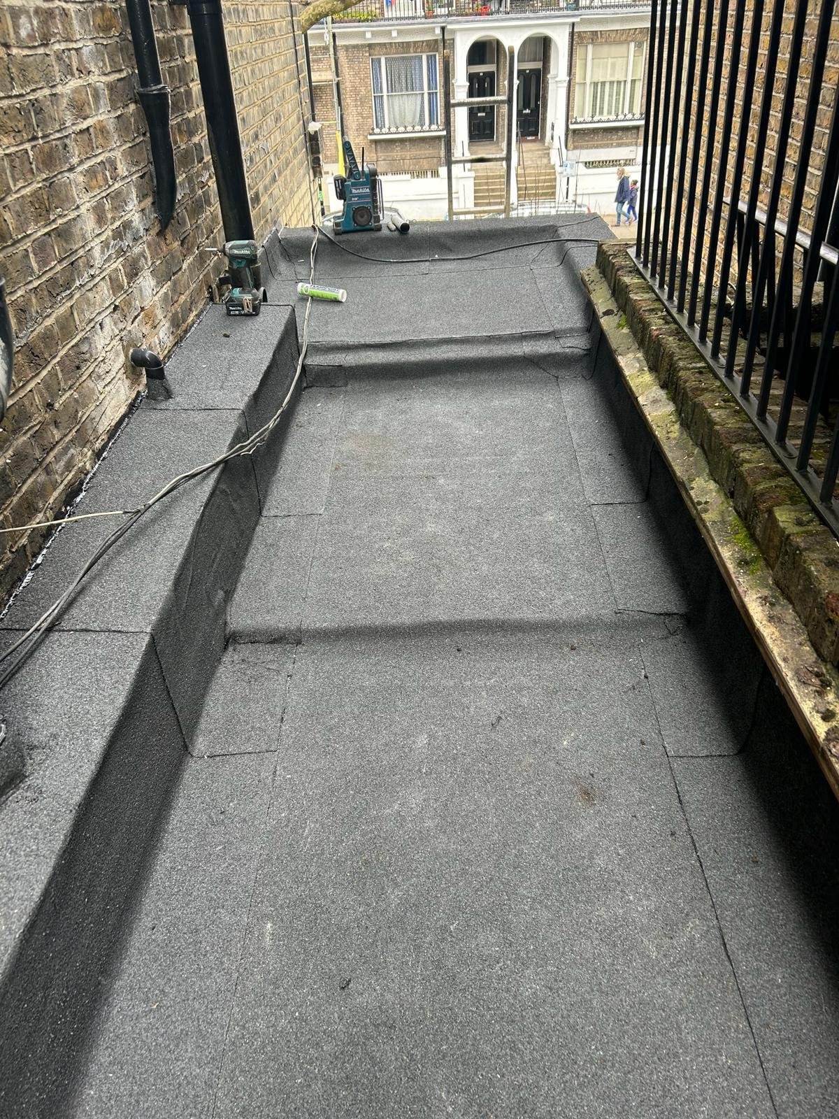A balcony with a new waterproof rubber roofing membrane, tools, and cords, overlooking a street with buildings and pedestrians.