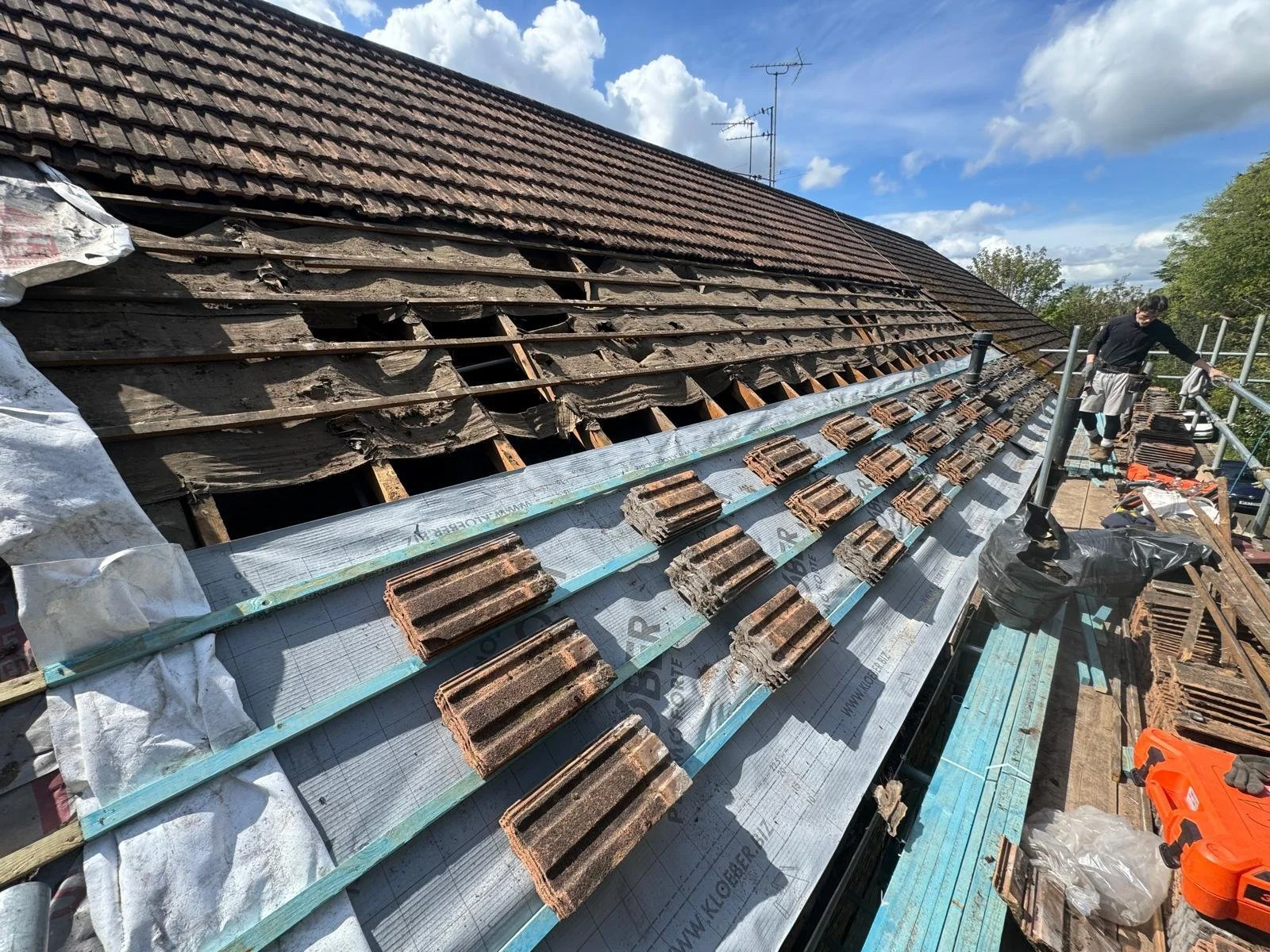 Roof being repaired, with wooden tiles removed and new tiles laid out, workers on scaffolding under cloudy sky.