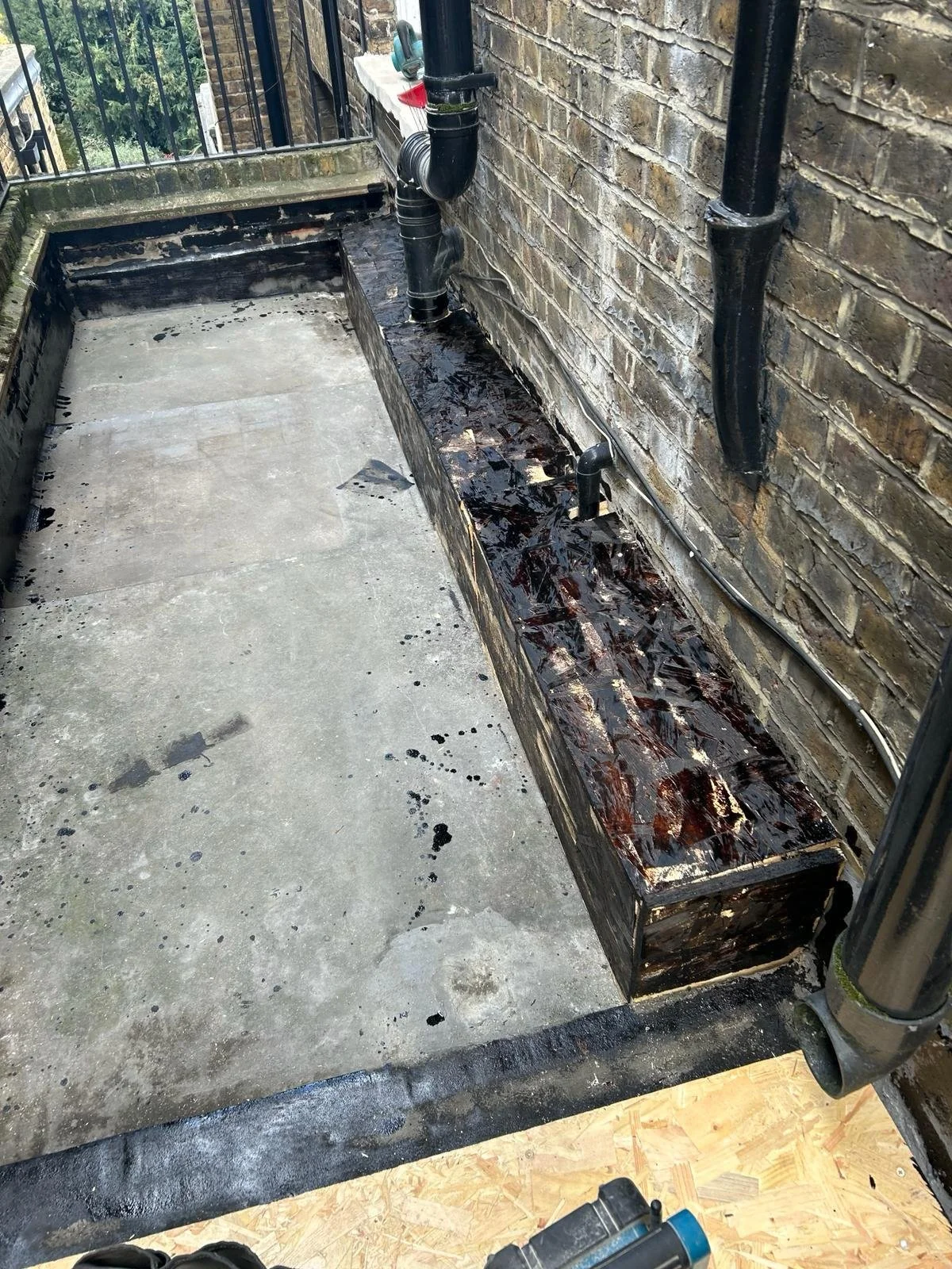 A balcony with a concrete floor and a brick wall with black pipes and a wood planter box. Some construction or renovation work is in progress.