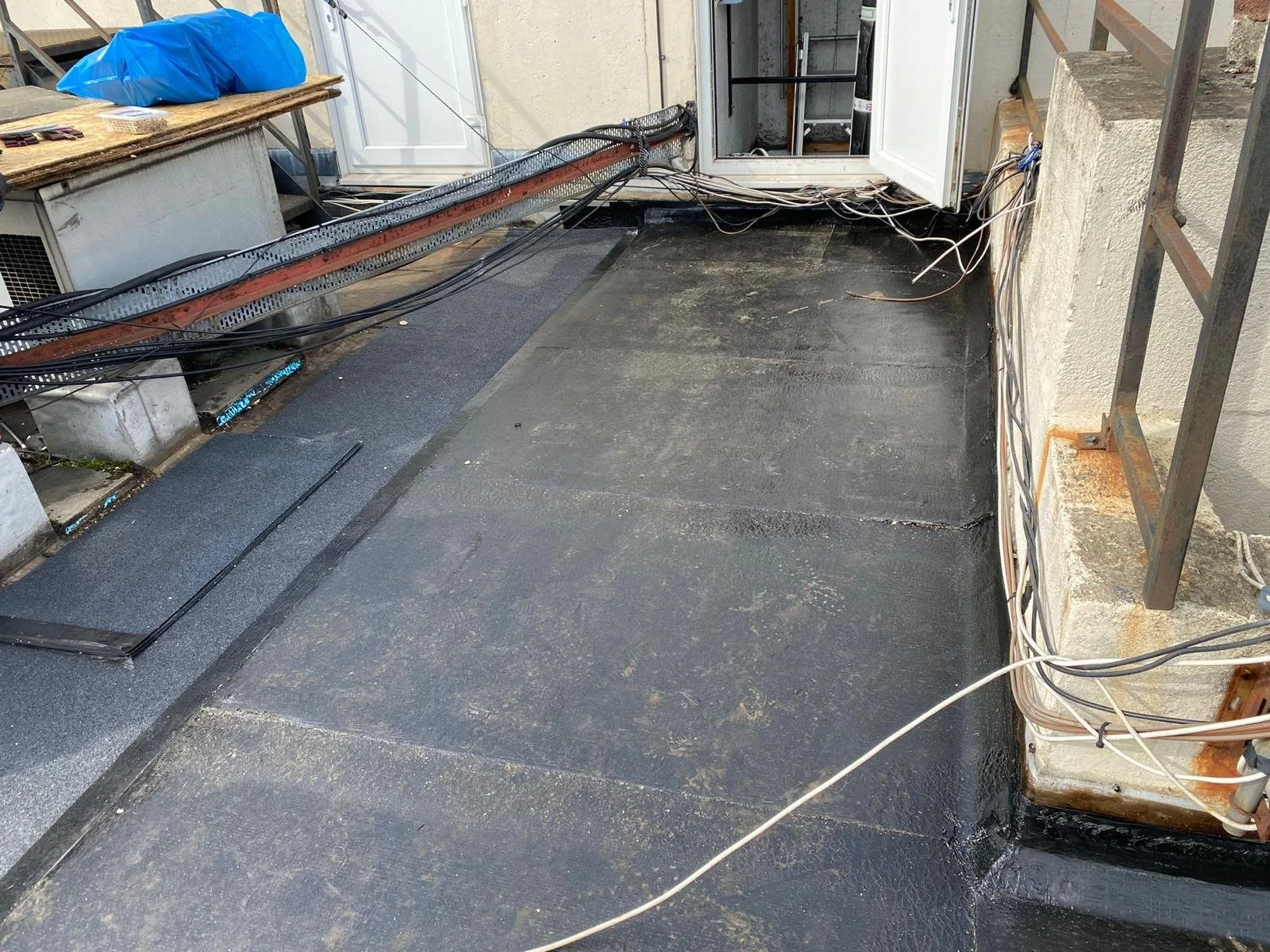 View of a rooftop with a black waterproof membrane, wires, and an open door leading inside a building.