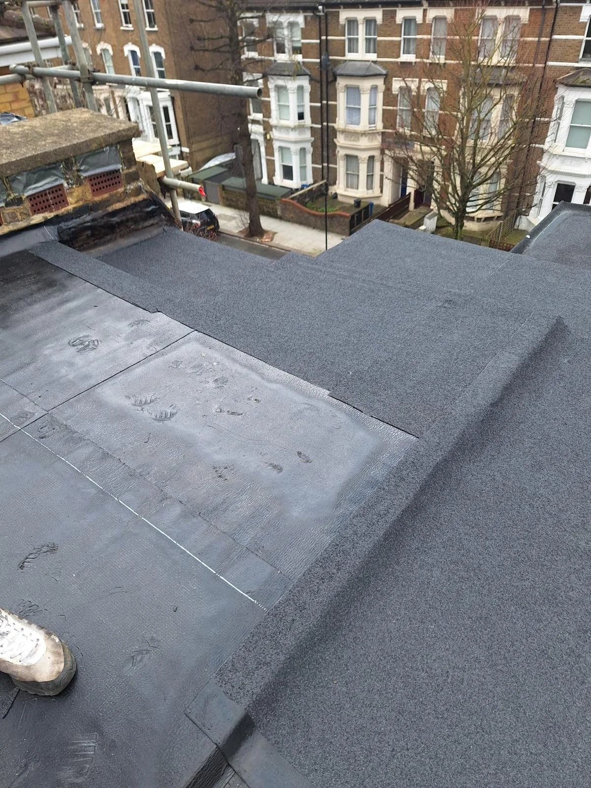 View of a rooftop with newly installed dark gray shingles, with a section of wet or freshly sealed surface, and a person wearing white shoes standing on the roof. In the background, there are residential buildings with bay windows and trees along the