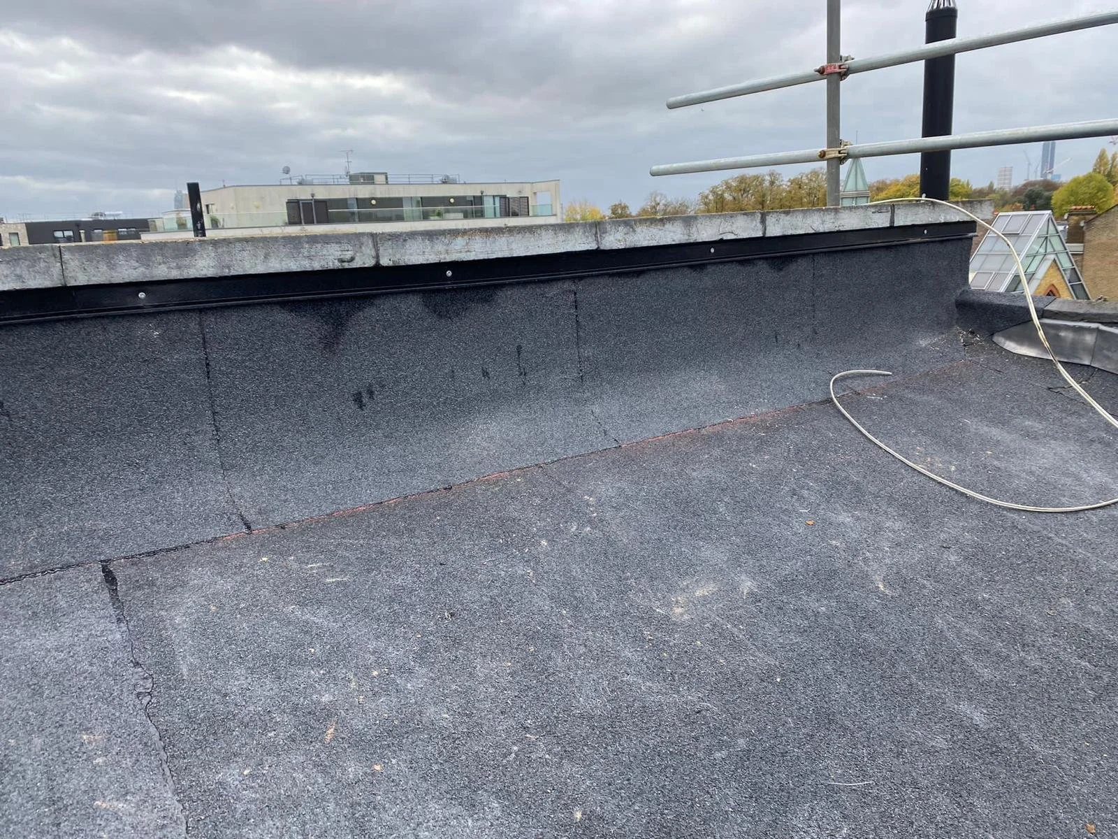 Flat rooftop with black waterproofing membrane, some gravel, metal railing, and an antenna, with city buildings and cloudy sky in the background.