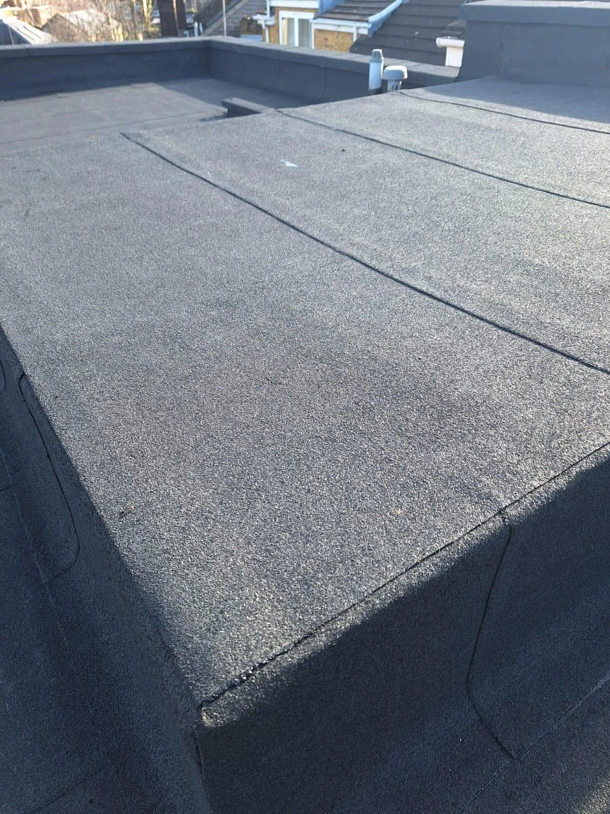 Close-up view of a flat, textured, dark gray asphalt roof with roof vents visible in the distance and roof shingles of neighboring houses in the background.