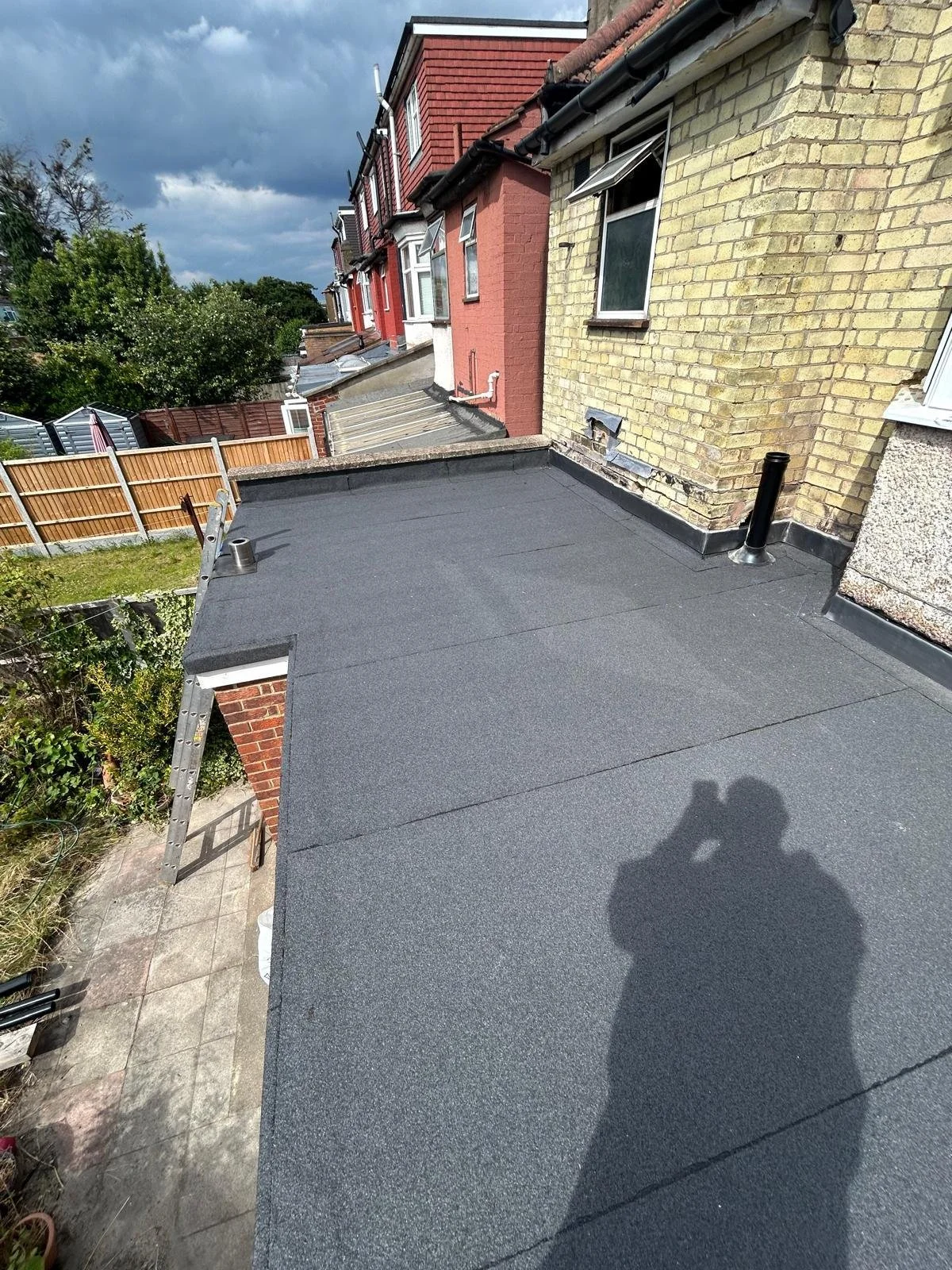 A flat rooftop with black roofing material, two black vents, and a ladder. Residential houses with brick and siding walls are visible, along with a fence and trees in the background. Shadows of a person taking the photo are on the roof.