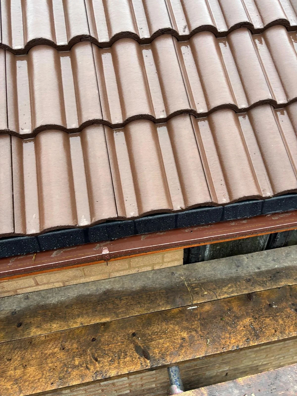 Close-up photo of a tiled roof with brown and terracotta-colored tiles, showing some black protective edging and part of a wooden structure underneath.