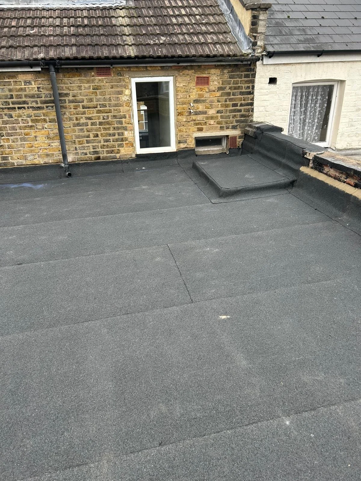 Flat rooftop with black roofing material, brick and white exterior walls, small window, and a window with lace curtains.