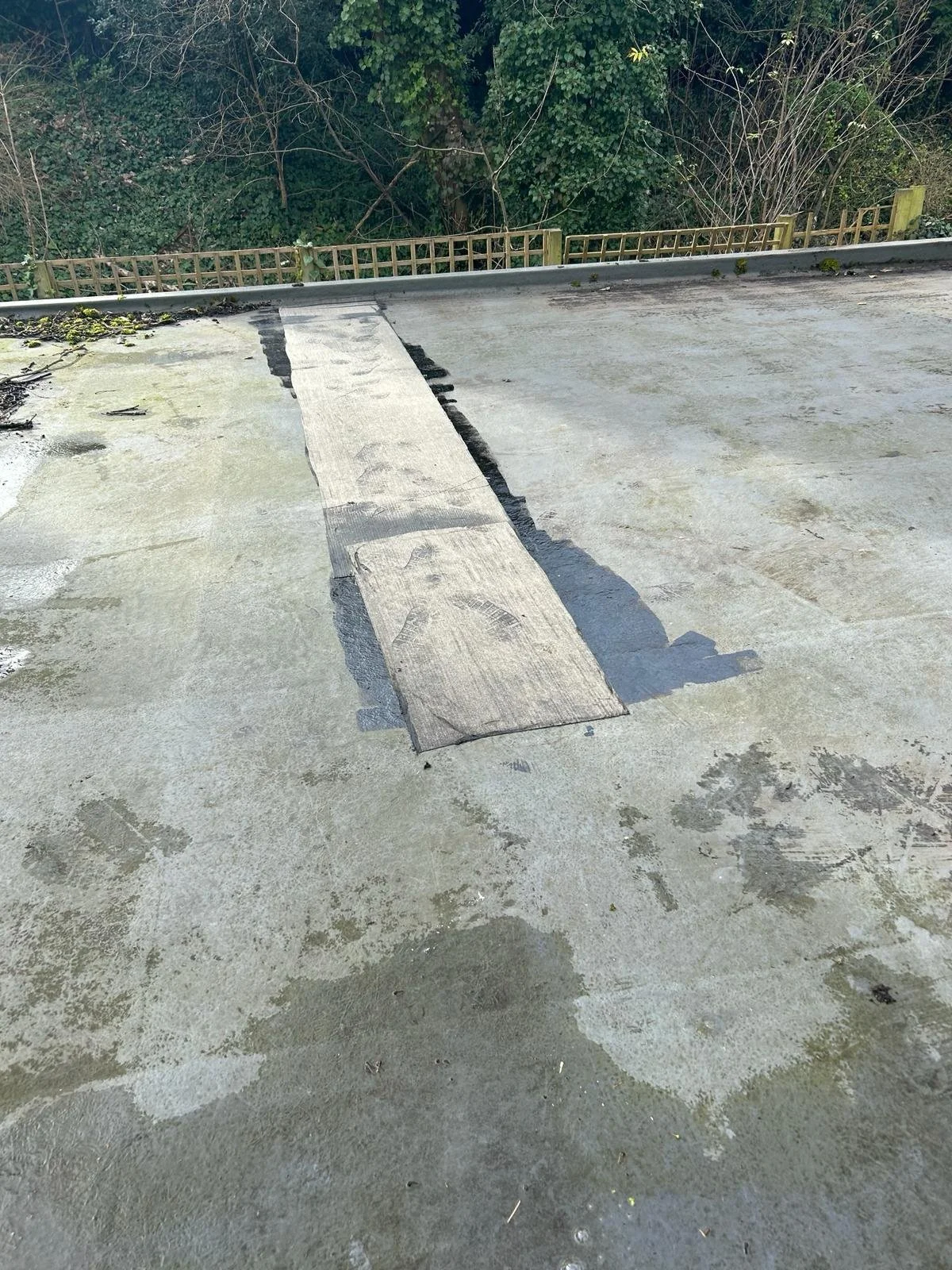 Flat rooftop surface with a wooden plank temporarily placed over a patch of black sealing or tar, surrounded by weathered concrete or asphalt, and a wooden fence with greenery in the background.