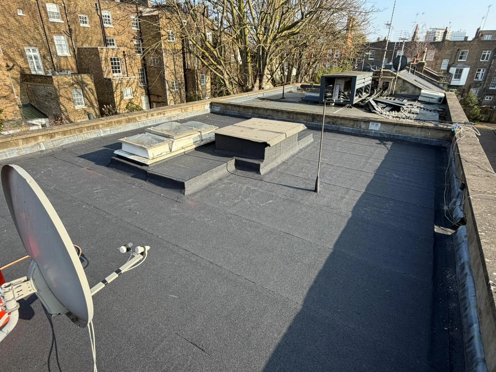 Rooftop with a satellite dish, two skylights, a chimney, and damaged equipment on the roof of a building, with brick apartment buildings and trees in the background.