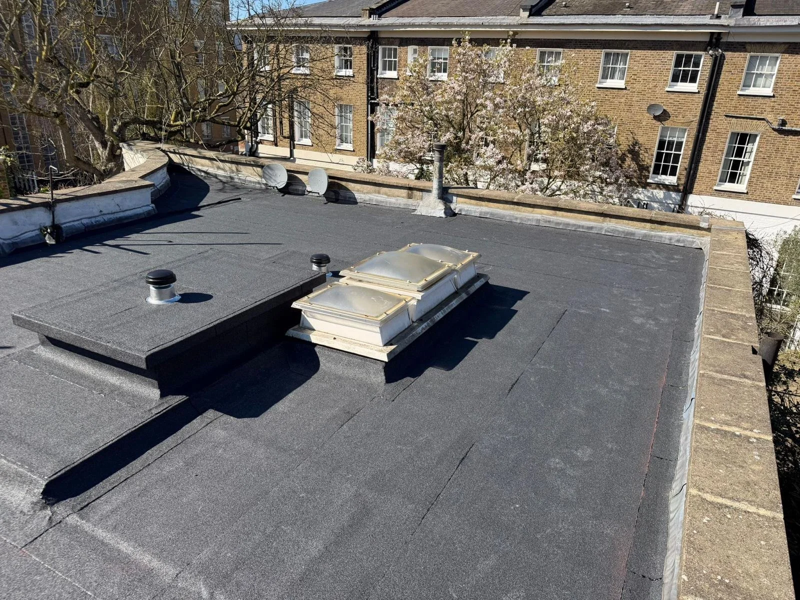 View of a flat rooftop with vents and skylights, surrounded by brick building walls and trees with blooming white flowers.
