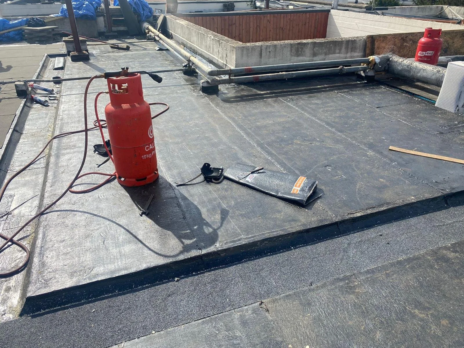 On a building roof, there are two red propane tanks, tools, pipes, and black roofing materials. The roof appears to be undergoing repairs or maintenance.