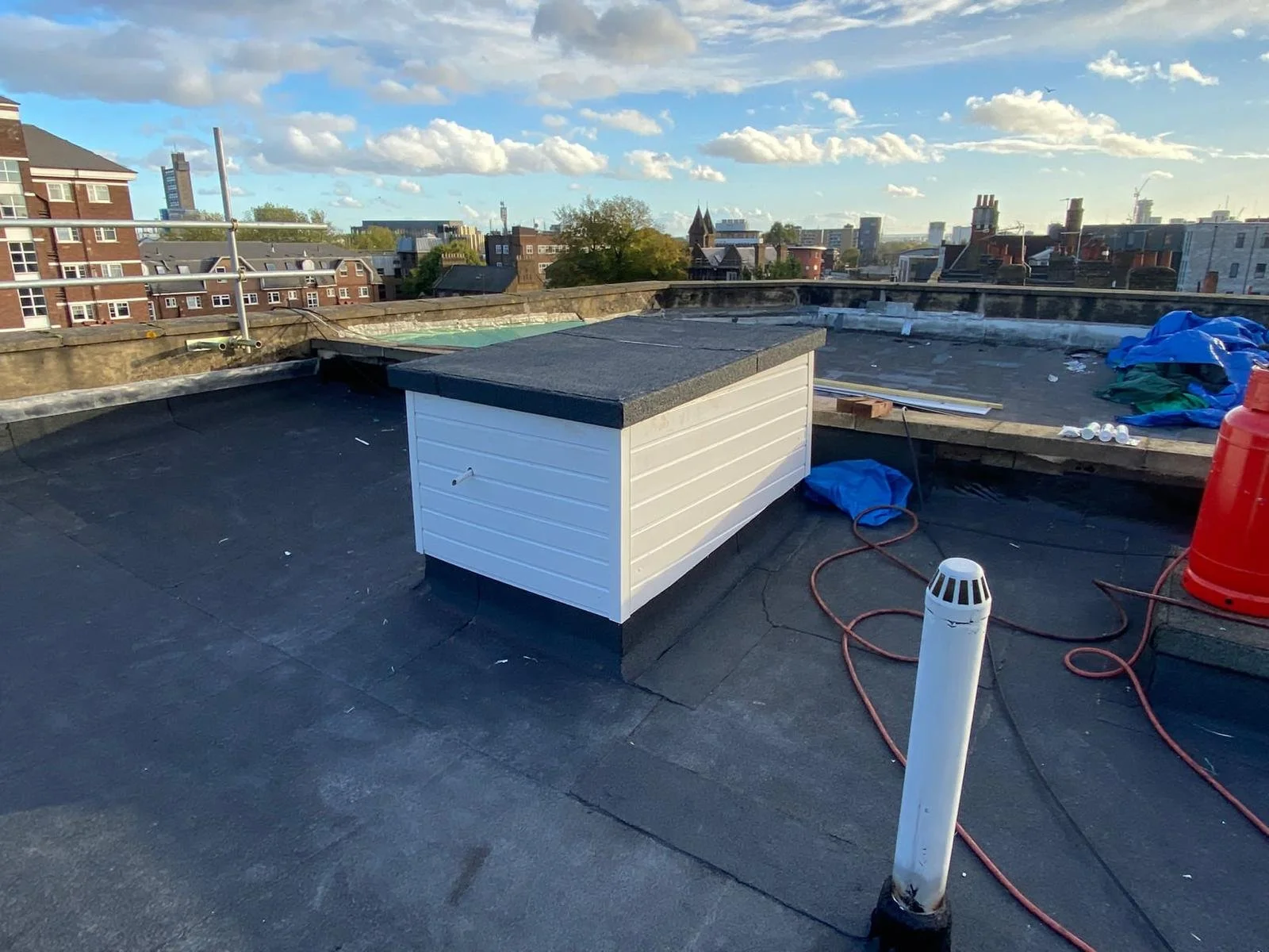 Rooftop with black roofing material, a small white wooden structure, umbrellas, tools, and cityscape with buildings under a partly cloudy sky.
