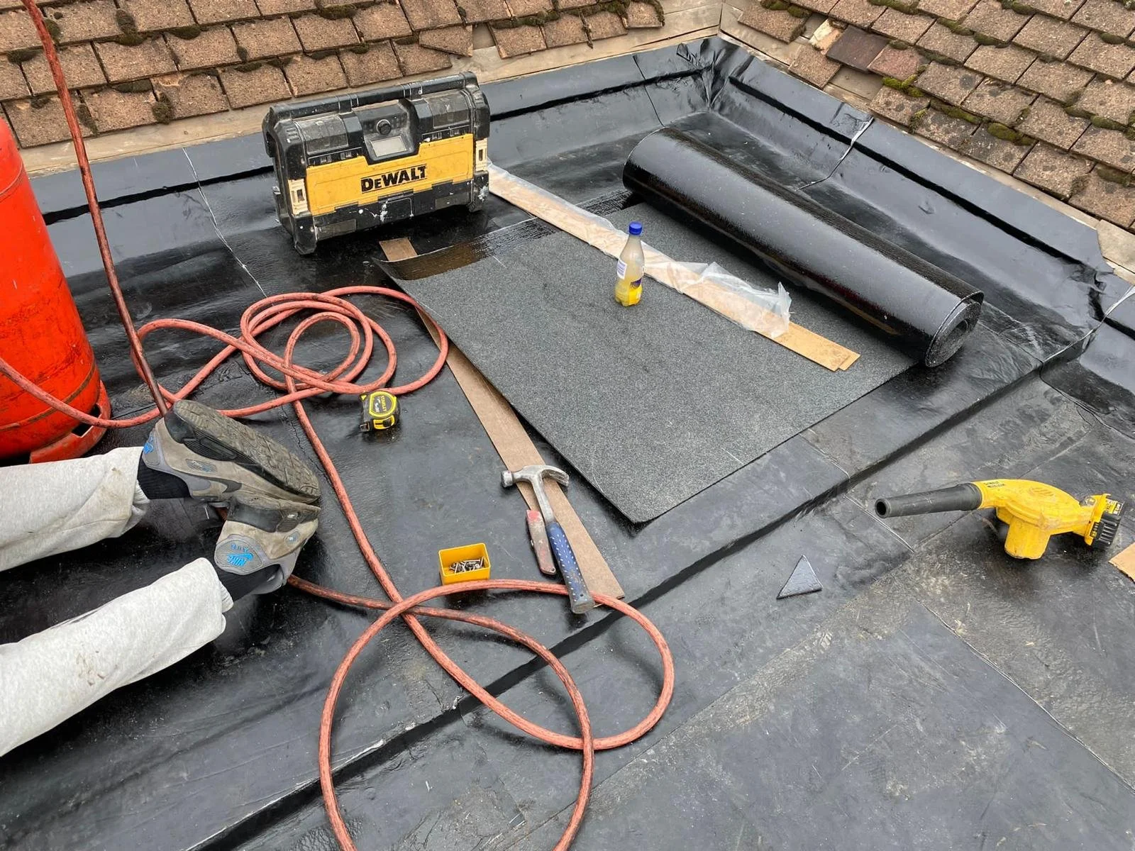 Construction worker installing roofing material on a flat roof, with tools and equipment nearby including a cordless drill, hammer, tape measure, and a DeWalt toolbox.