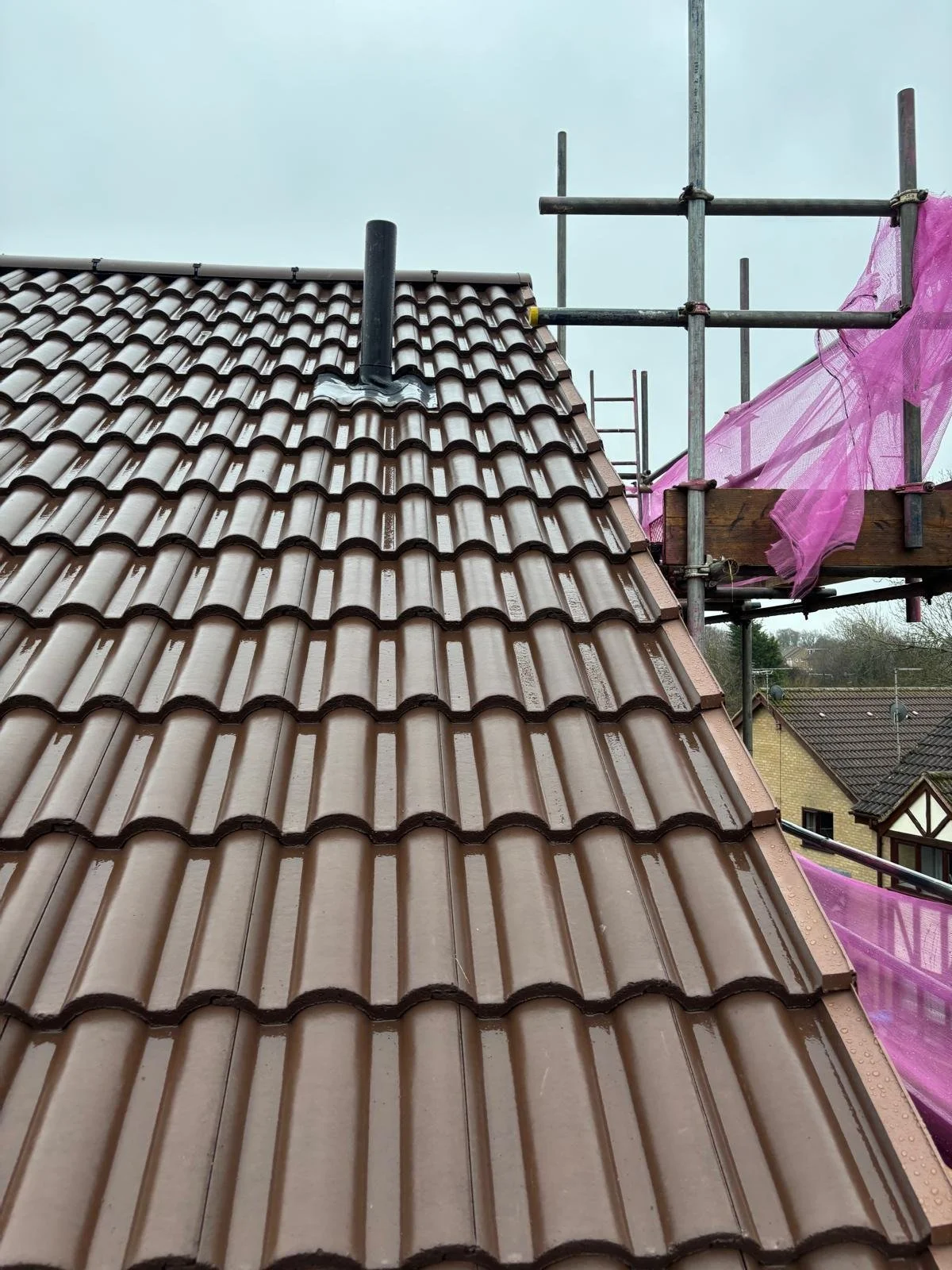 Photo of a brown tiled roof with scaffolding on the side, pink construction netting, and a cloudy sky in the background.