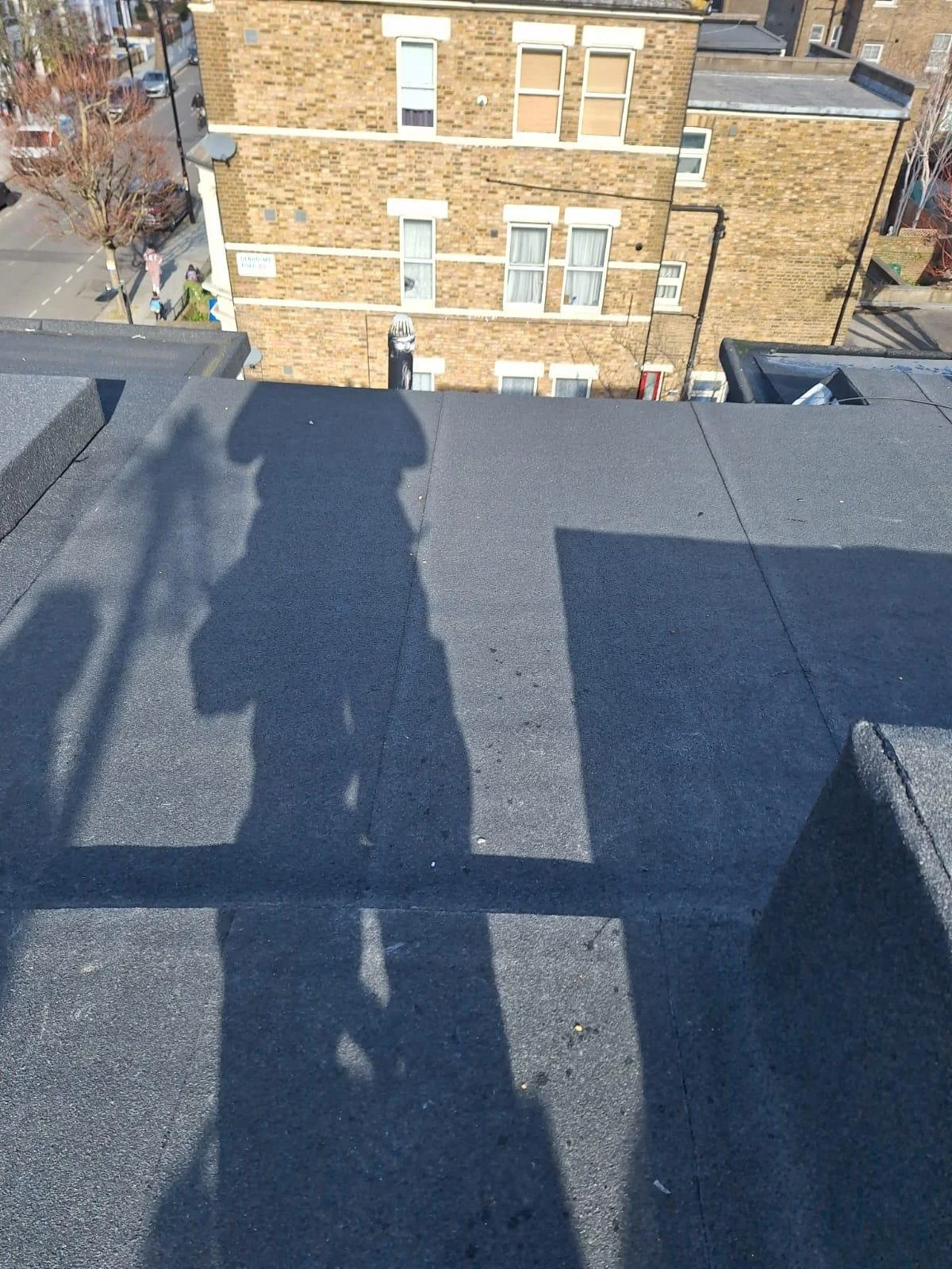 Shadow of a person taking a photo on a flat rooftop with a brick building and street below in the background.