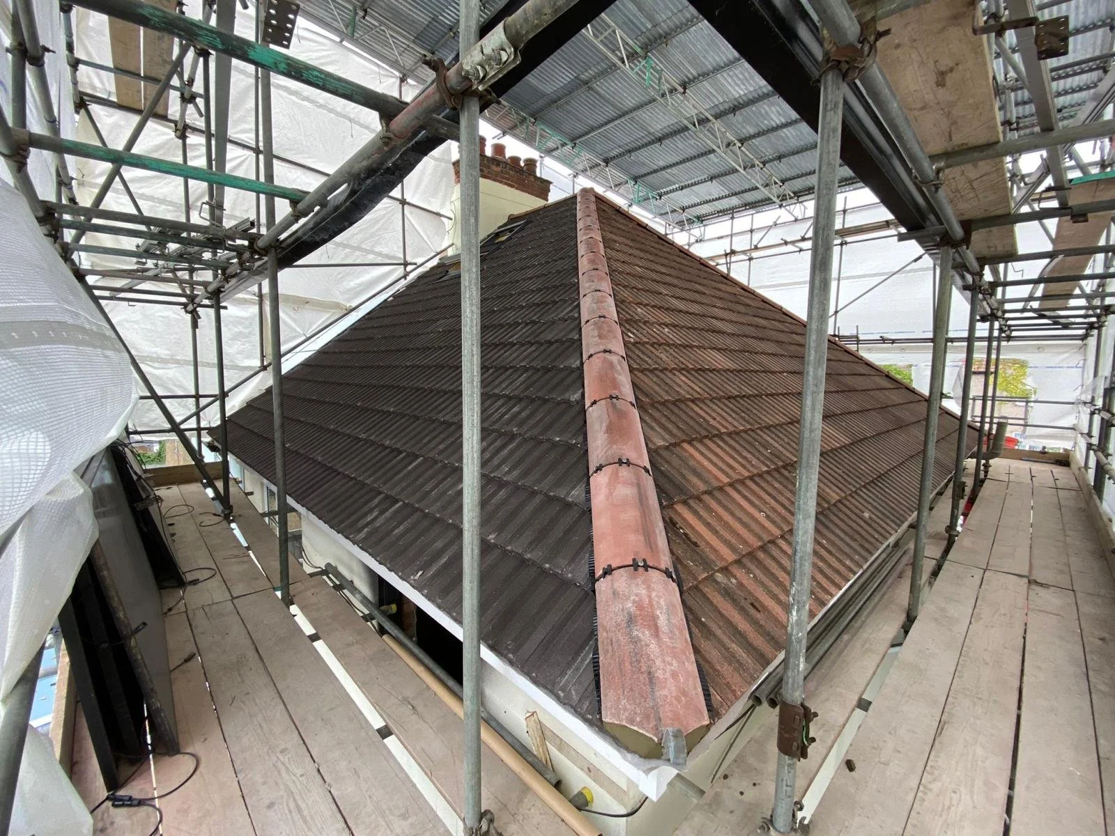 View of a sloped tiled roof of a building under construction or renovation, surrounded by scaffolding, with visible scaffolding poles, safety barriers, and building materials.