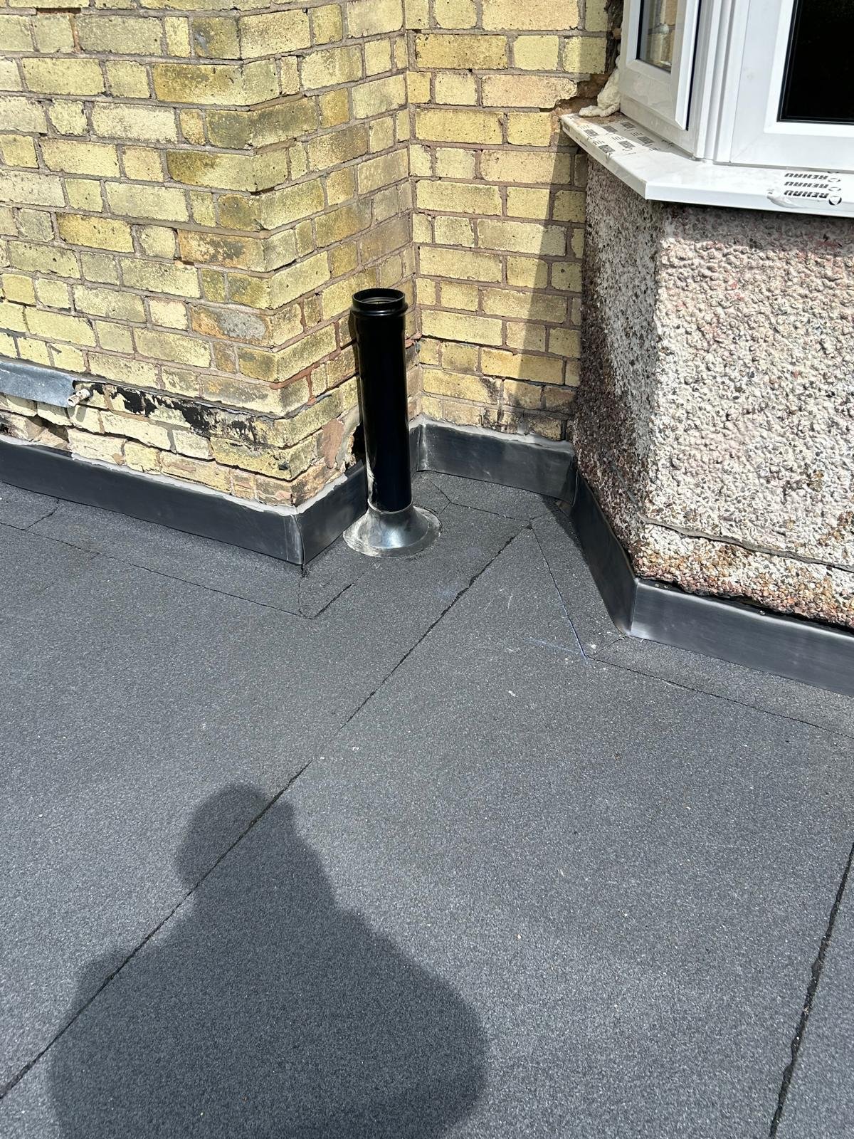 Black pipe protruding from the roof near a brick wall and a window with a white frame. The roof surface is covered with gray waterproof material.