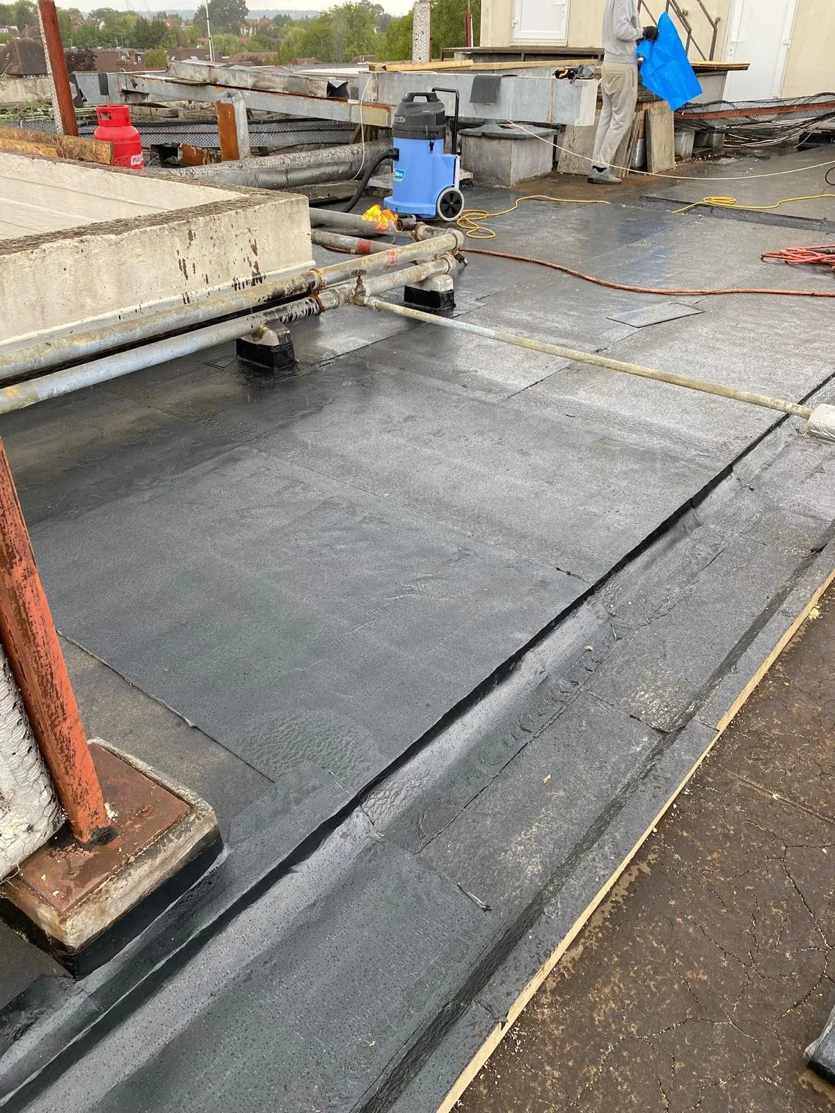 Flat roof with black sealant coating, exposed pipes, and construction workers in the background working on the building's edge.