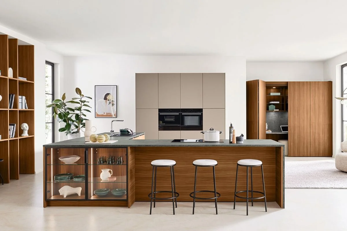 Cuisine moderne avec île centrale en bois clair, bar avec trois tabourets blancs, étagère en bois à gauche, meuble encastré blanc avec appareils électroménagers, plantes vertes, décoration minimaliste.