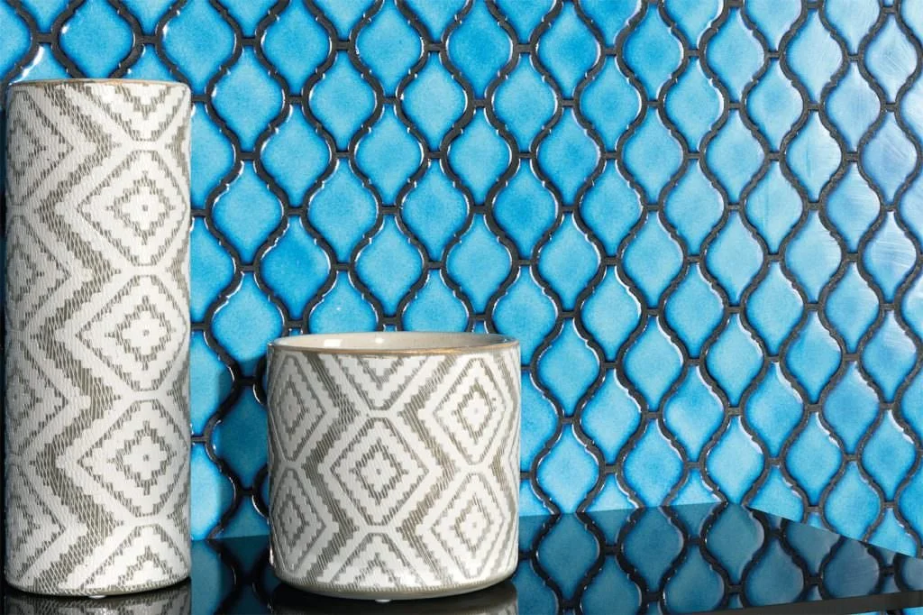 Deux rouleaux de papier toilette et un pot à crayons avec un motif géométrique blanc et gris, placés devant un fond en mosaïque de carreaux bleus avec un motif en forme de croix noire.