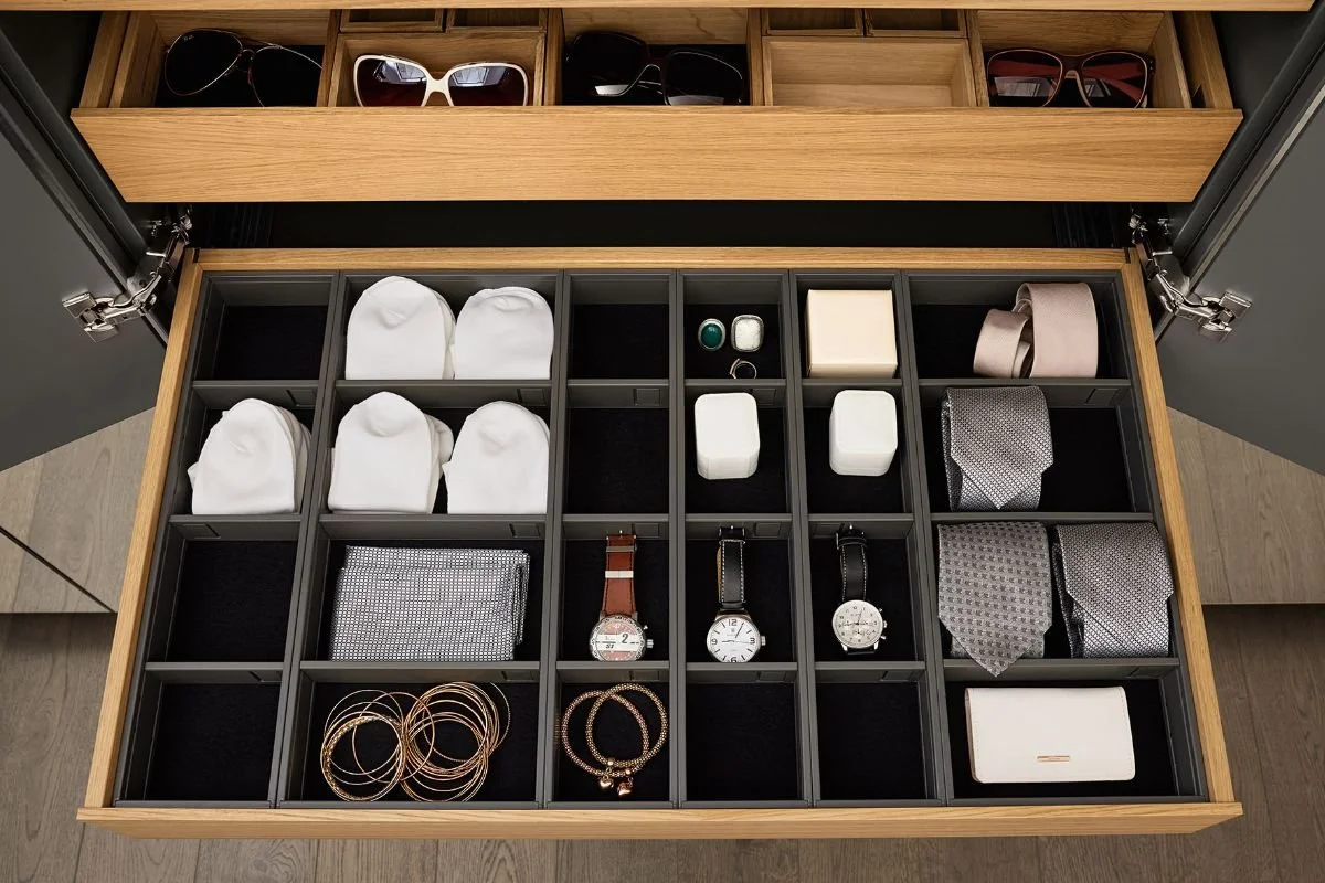 Un tiroir de coffre à accessories avec des montres, des boutons de manchette, des ceintures, des cravates, des bijoux et des étuis à cigare. Au-dessus, un tiroir avec des lunettes de soleil.