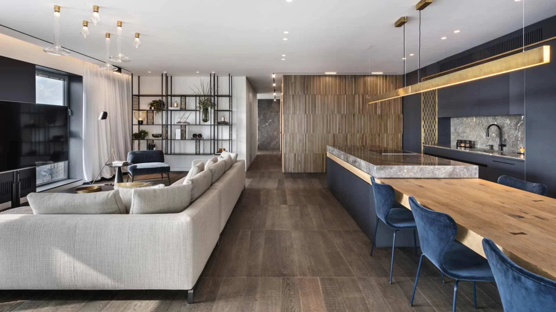Intérieur moderne d'un salon et cuisine avec canapé beige, étagères métalliques, plan de travail en marbre, chaises bleues en velours, et éclairage suspendu doré.