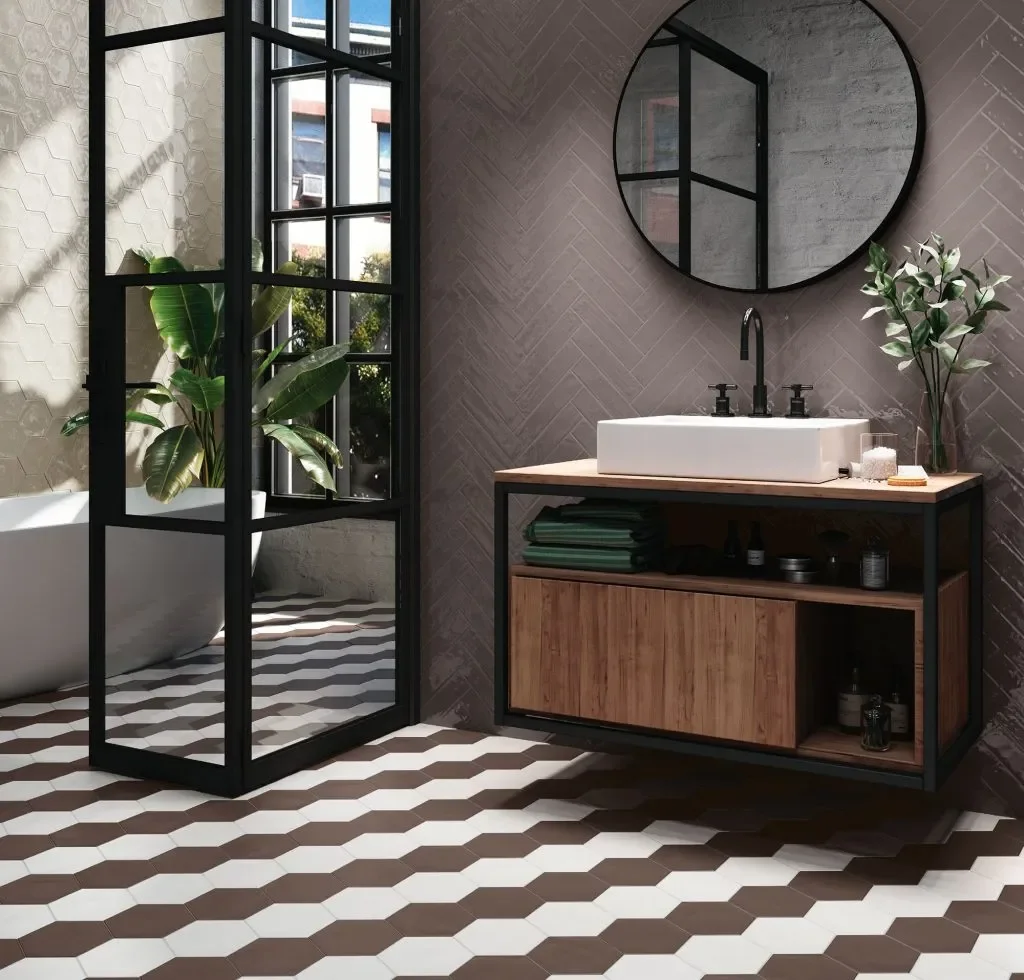 Une salle de bain moderne avec un évier blanc sur un meuble en bois, un miroir rond noir, une plante verte, et une douche séparée avec une paroi en verre noir, et un mur en tuiles grises, avec un sol en carrelage hexagonal blanc et marron.