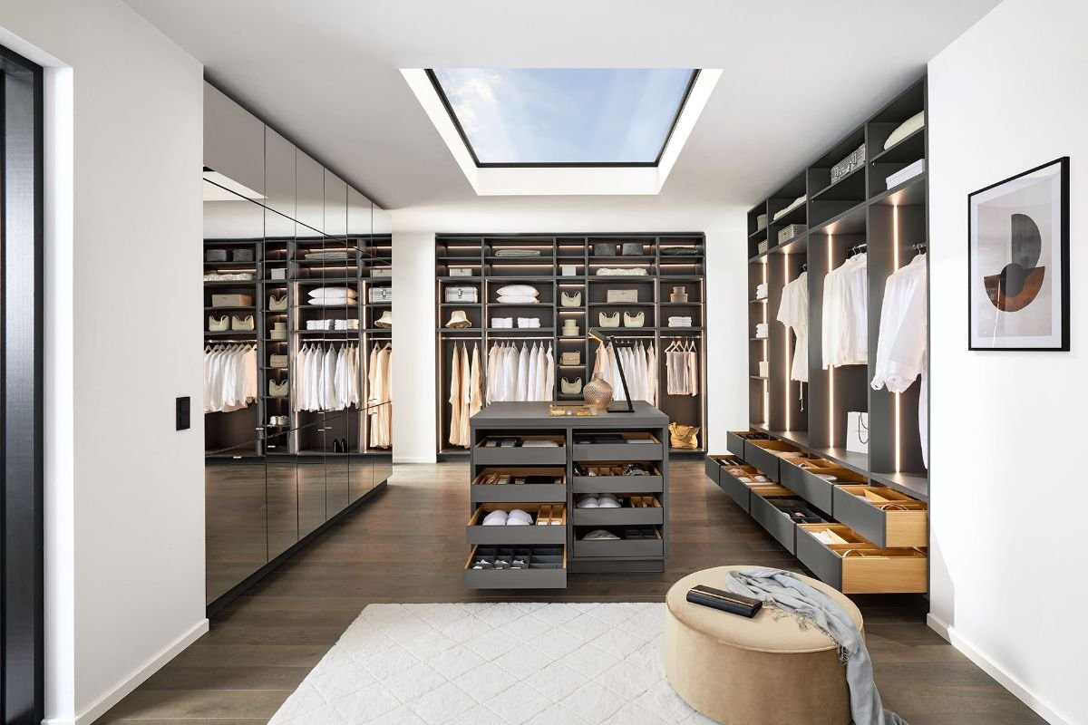 Une chambre de dressing moderne avec des étagères en bois foncé, des tiroirs, des vêtements blancs suspendus, un trou de toit avec vue sur le ciel, un tapis blanc, un ottoman beige et un tableau abstrait au mur.