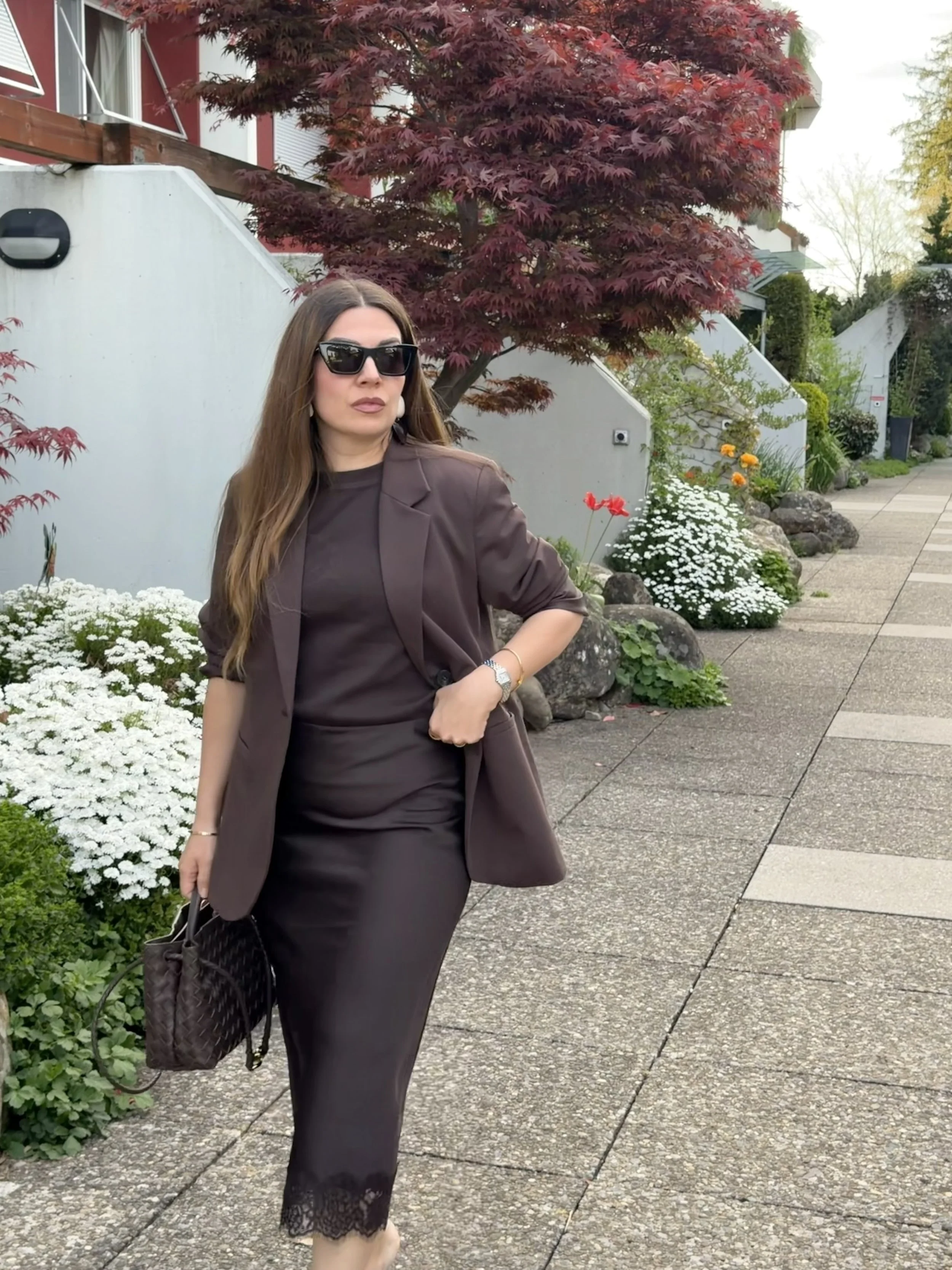 A woman with long brown hair wearing black sunglasses, a dark brown blazer, and a black dress with lace at the bottom, stands on a sidewalk next to a garden with white and orange flowers, and a large red-leafed tree in the background.