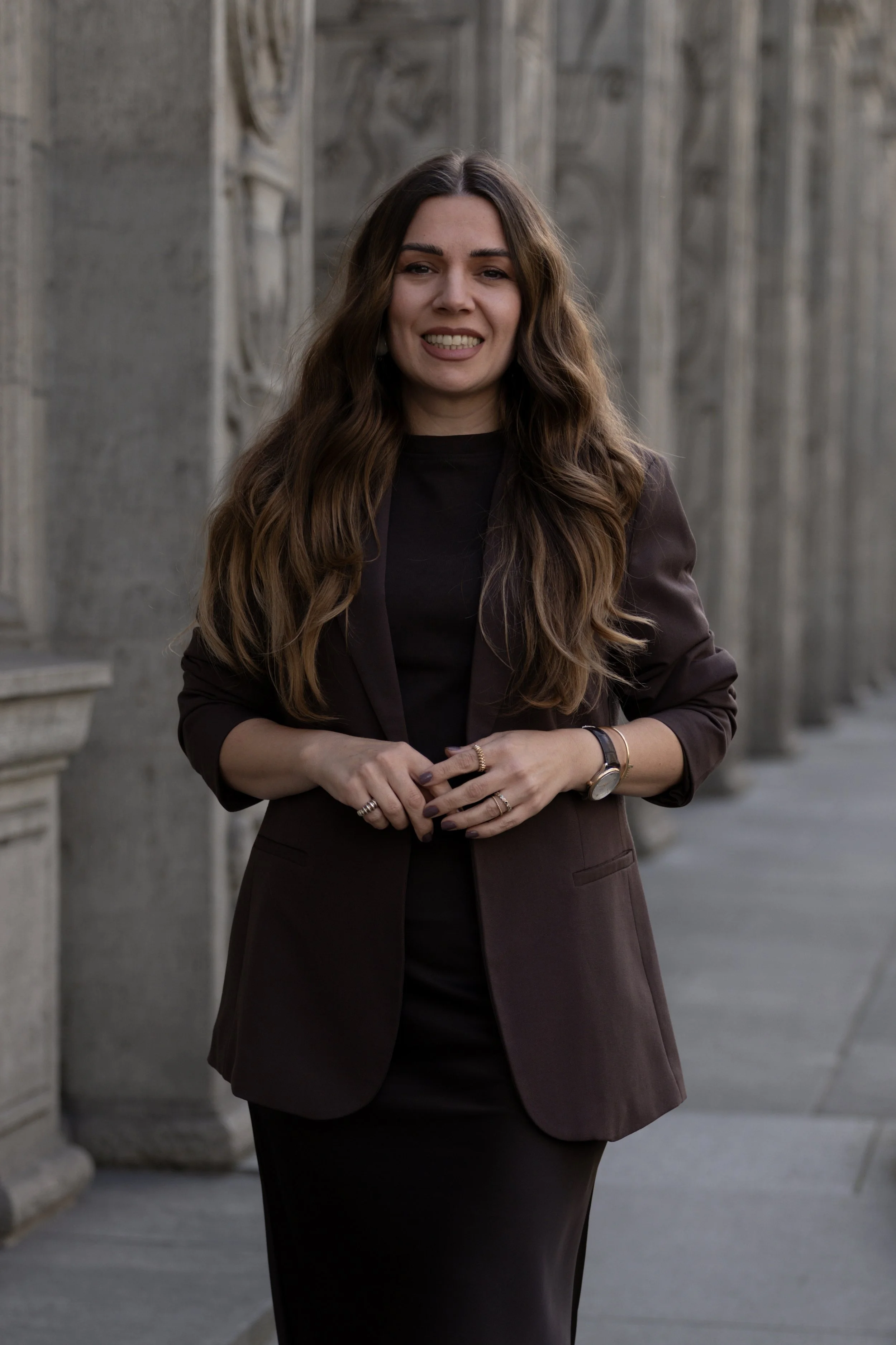 A woman with long wavy brown hair, wearing a dark blazer and skirt, standing outdoors in front of a gray stone building with carved architectural details, smiling with her hands clasped together.