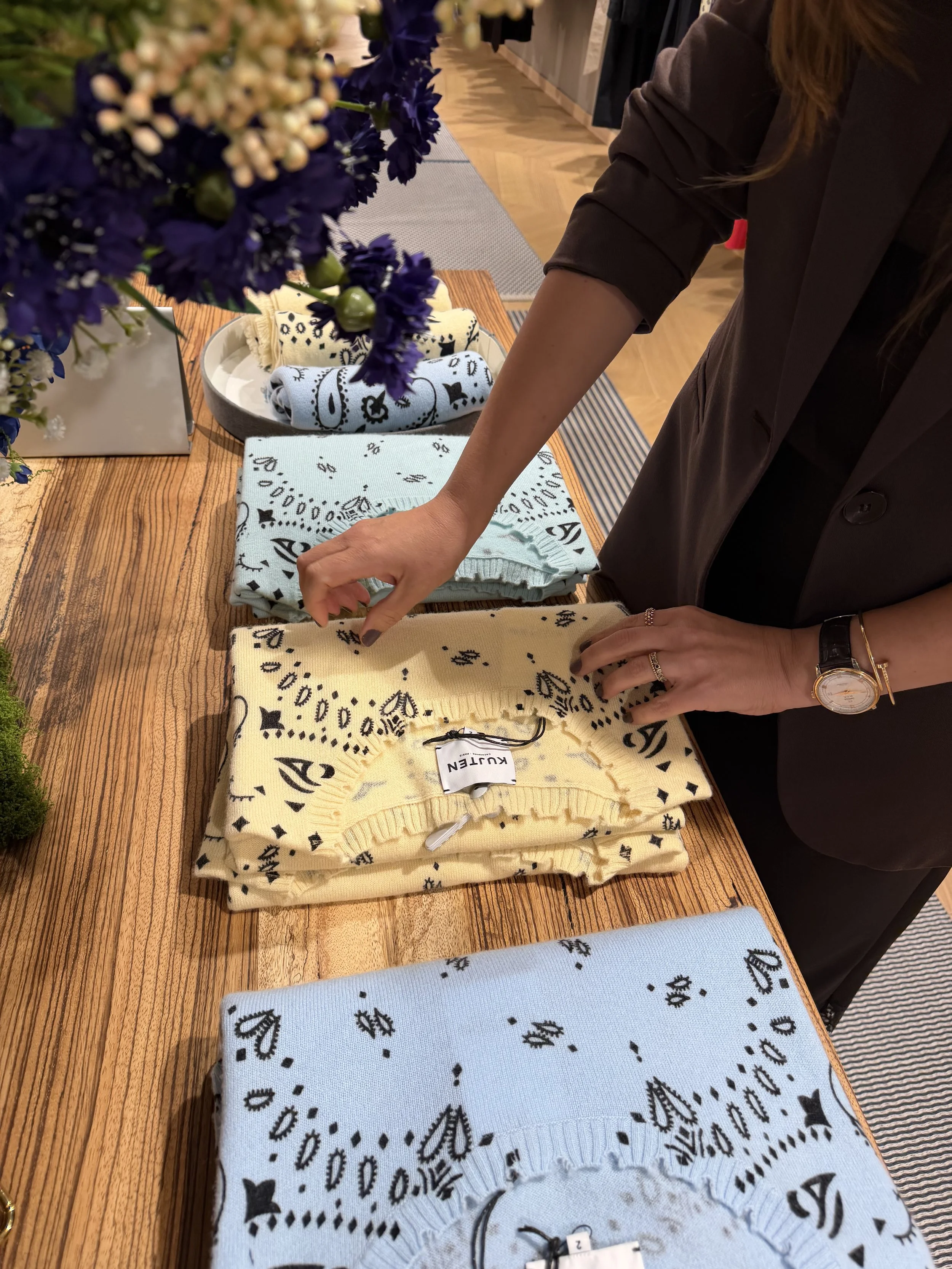 Display of folded knit scarves or sweaters with black duct tape ribbon tags, arranged on a wooden table, with a person partially reaching for the items, in a retail store environment.
