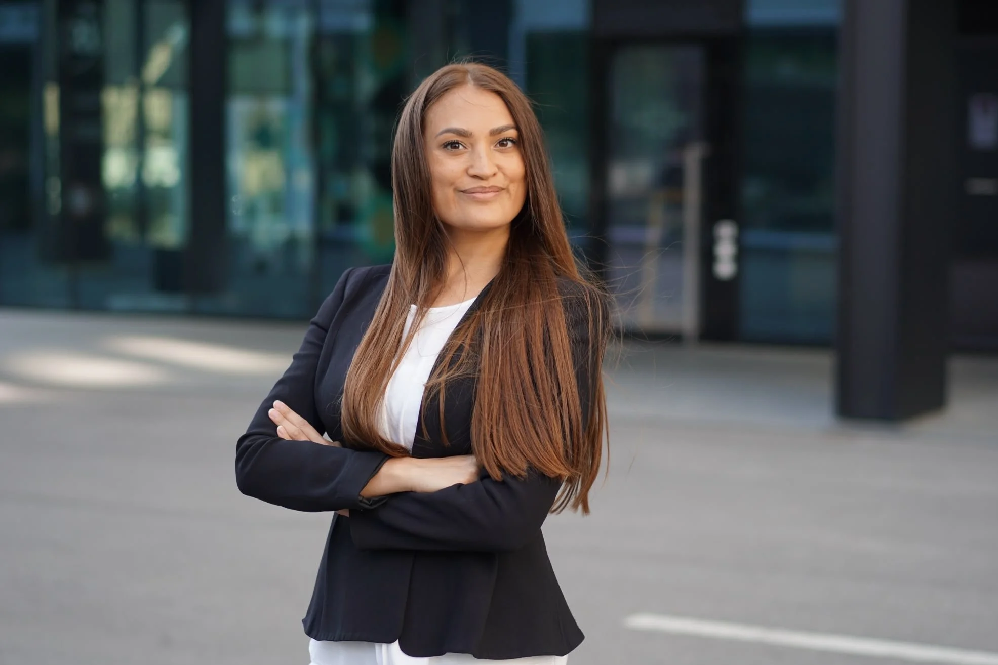 A confident woman with long brown hair, dressed in a black blazer and white shirt, standing outside with her arms crossed near a modern glass building.