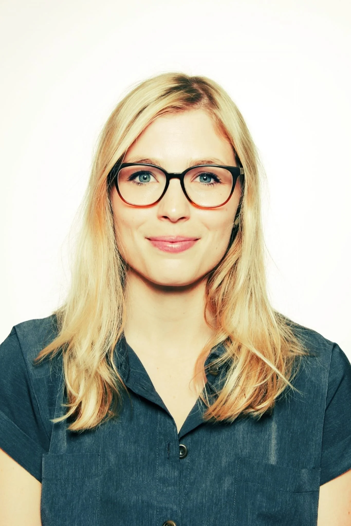 A woman with blonde hair, wearing glasses and a dark button-up shirt, smiling in front of a plain white background.