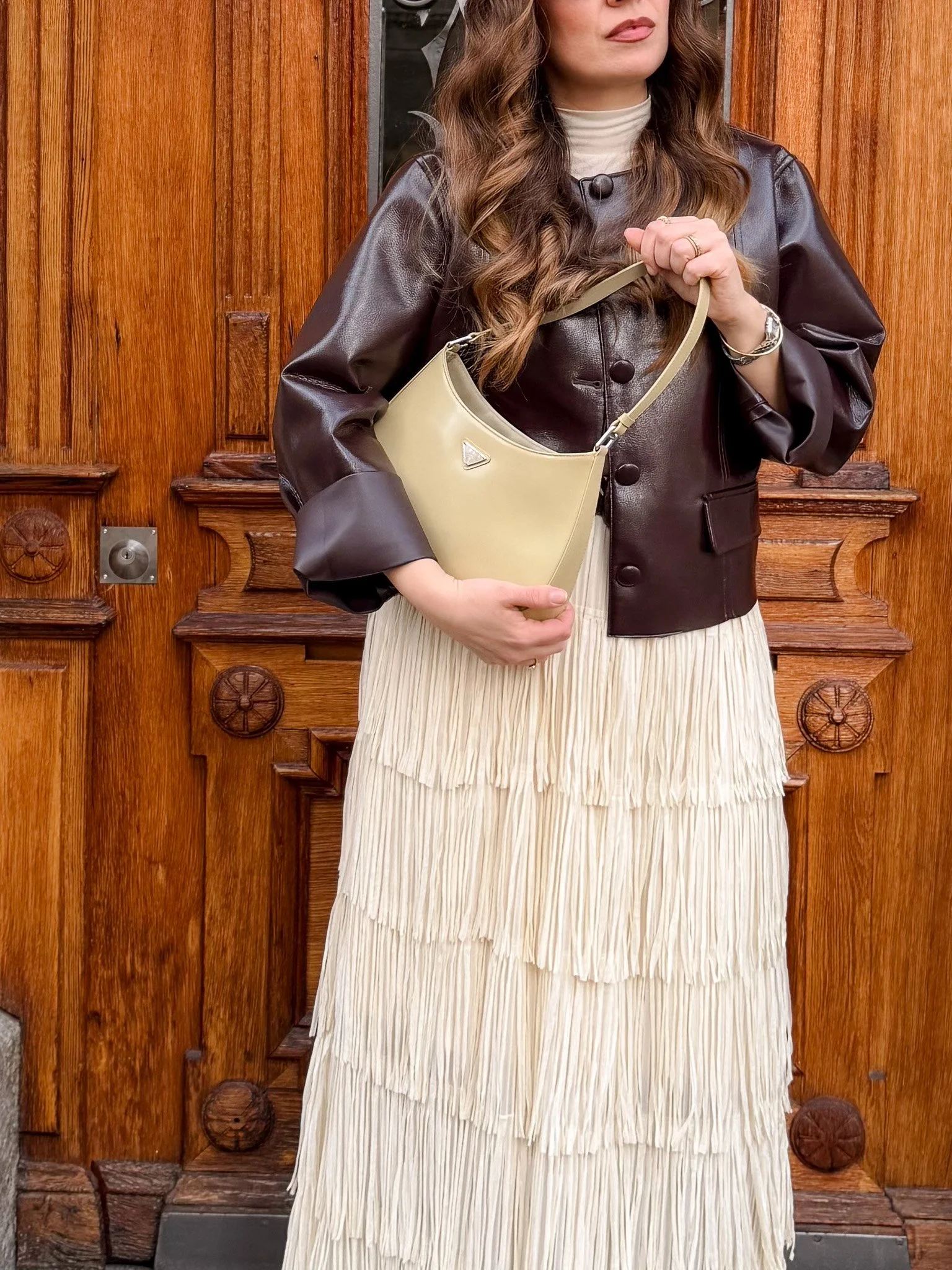 Woman with long wavy brown hair wearing a cream pleated dress, a shiny black leather jacket, holding a beige handbag, standing in front of a wooden door.