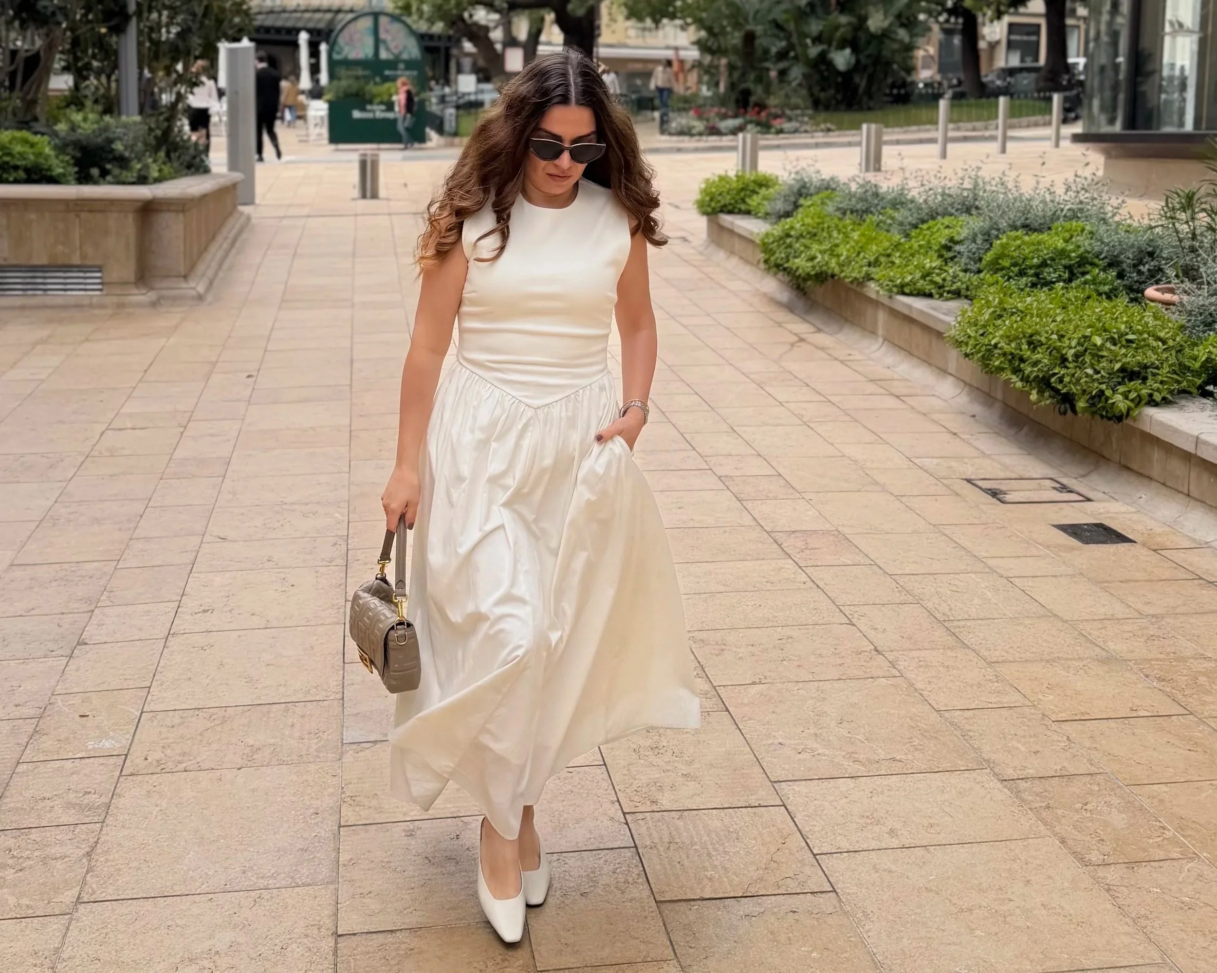 Woman in a white dress and white boots walking outdoors on a paved surface, wearing sunglasses and carrying a small handbag.