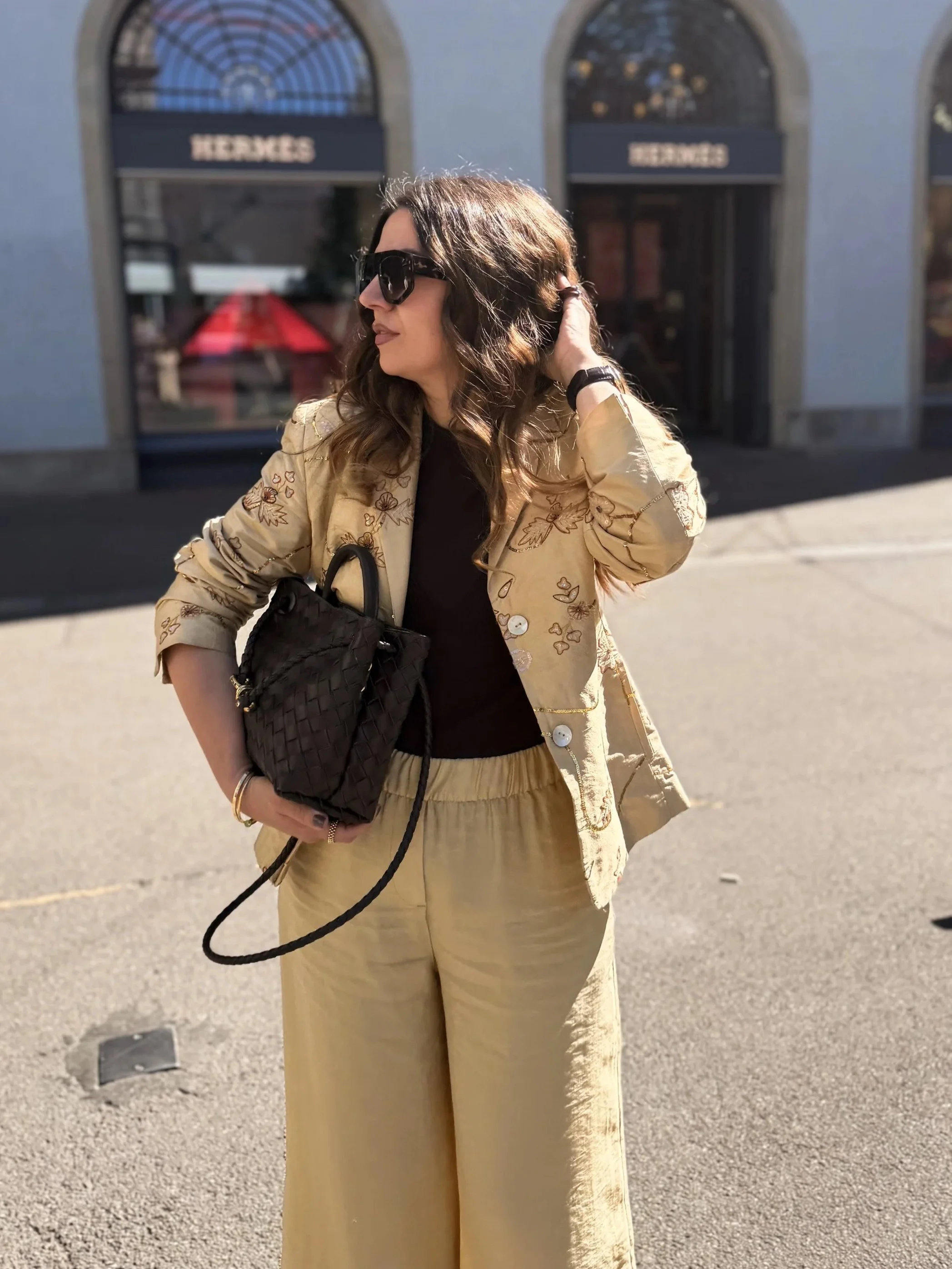 A woman standing outdoors, wearing large sunglasses, a beige embroidered jacket, and beige pants, holding a black quilted handbag with a long strap, with a building with arched windows and the word 'HERMES' in the background.