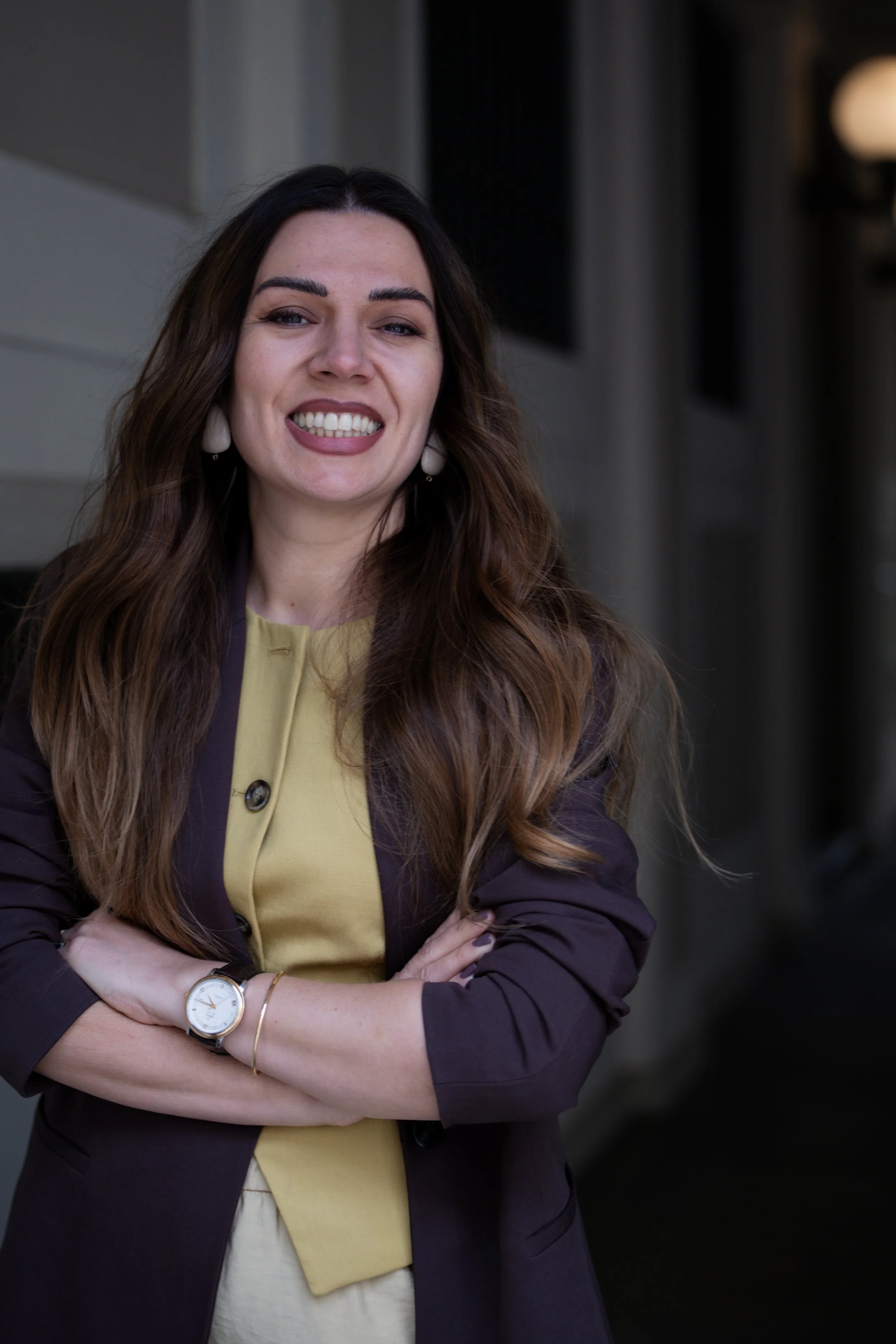 A woman with long brown hair smiling, crossing her arms, wearing a yellow top and a dark blazer, standing outdoors near a building.