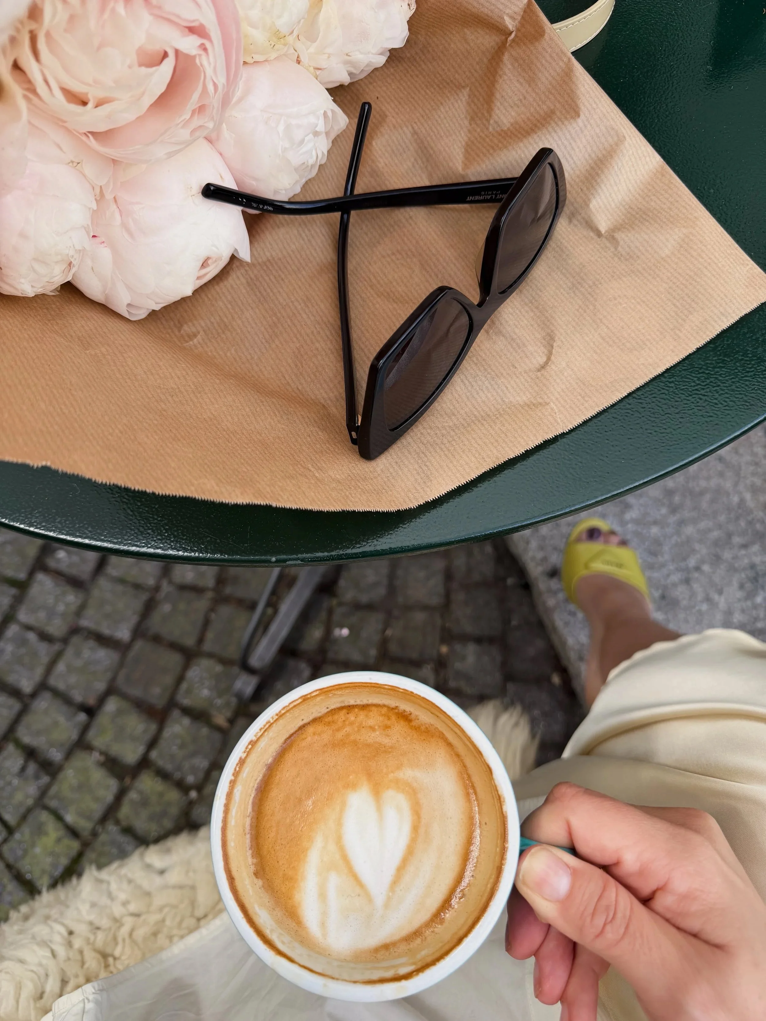 A person holding a cup of latte with foam art, on a table with a bouquet of pink and white flowers, a cup of rice, and sunglasses.