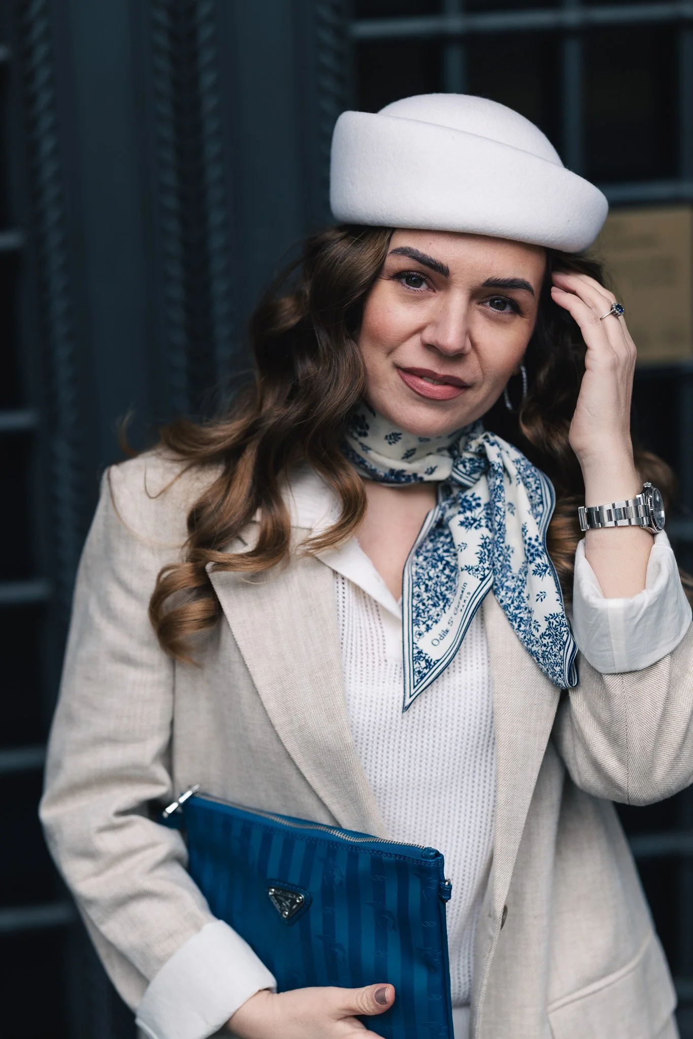 A woman with curly brown hair wearing a white hat, beige blazer, white shirt, and a blue and white scarf, holding a blue clutch purse and touching her forehead, standing in front of a dark background.