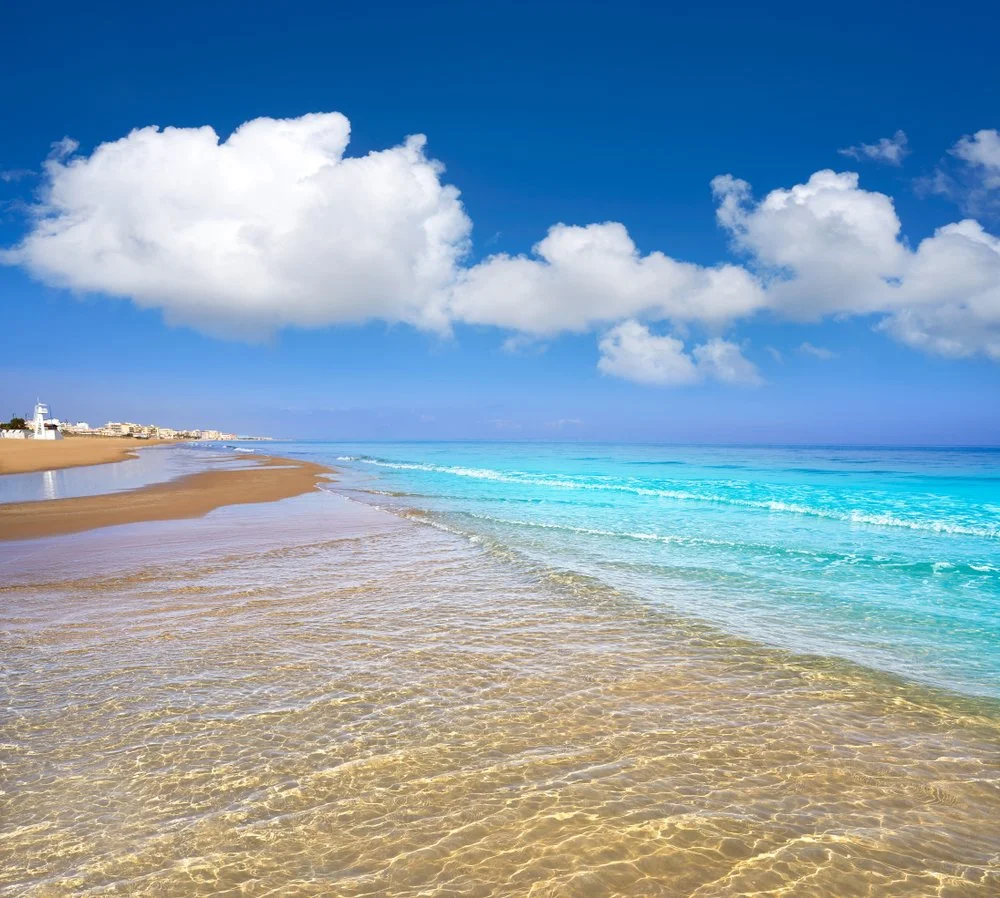 Strand met helder zeewater, witte wolken in een blauwe lucht, en gebouwen aan de horizon.
