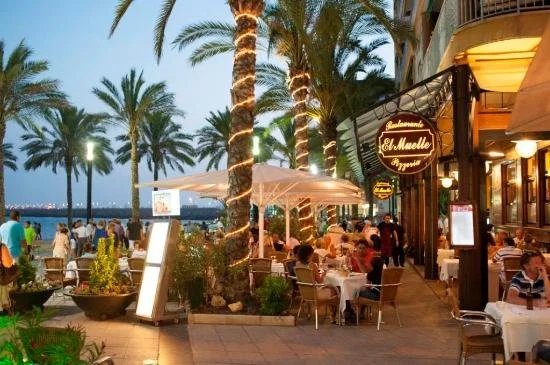 Gezellig restaurant met mensen die dineren, palmbomen met lichtjes, uitzicht op de zee in de avond.