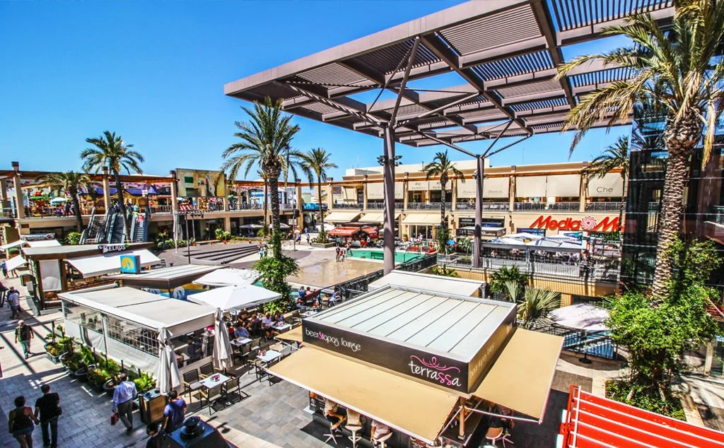 Uitgaansgebied met restaurants, winkels en palmboom rondom, mensen sitting en wandelen in een markt of winkelcentrum, zonnige dag.