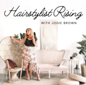 A woman standing in a bright, stylish living room with a white sofa and plants, holding a hairbrush or makeup brush, for a show called 'Hairstylist Rising with Jodie Brown'.