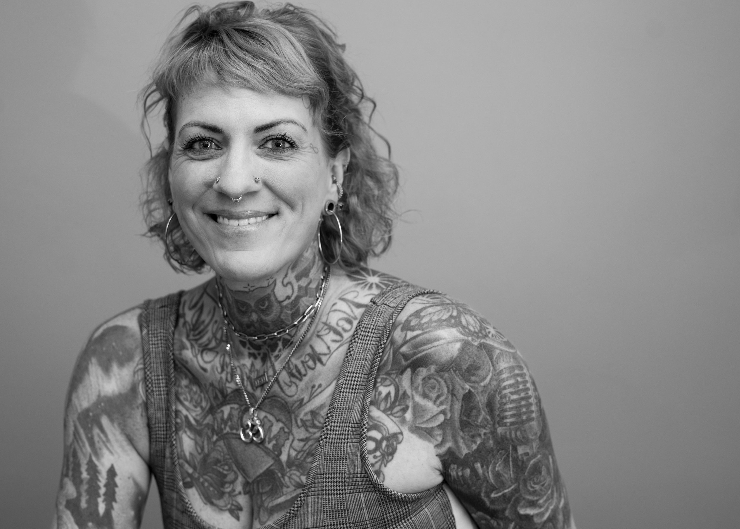 Black and white portrait of a smiling woman with short, curly hair, multiple facial piercings, and extensive arm and neck tattoos, wearing a sleeveless checkered top and layered necklaces.