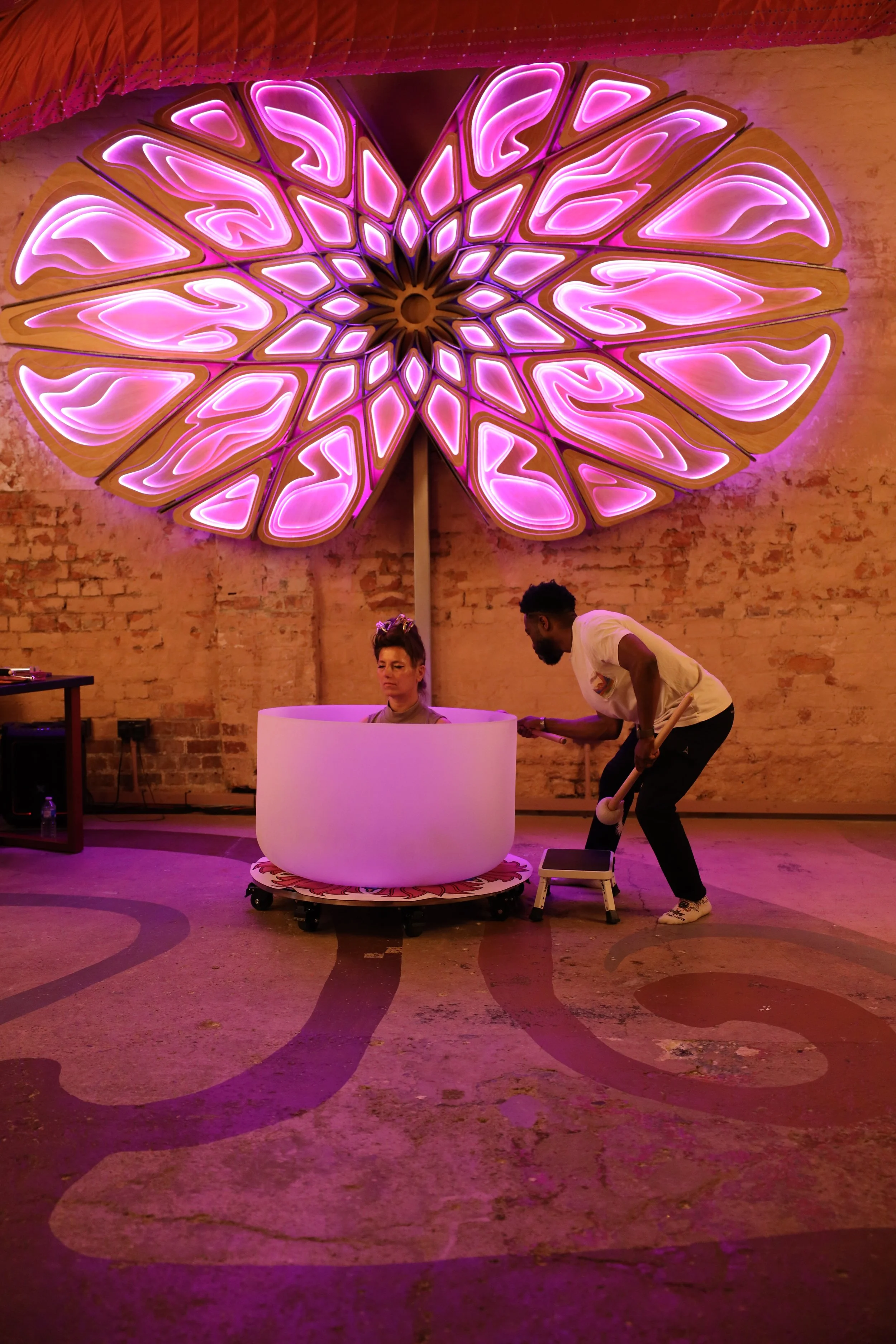 A woman is seated inside a large white drum with glowing pink and purple decorative lights resembling flower petals on the wall behind her. A man stands beside the drum, holding a mallet, preparing to strike it. The setting has an industrial background with exposed brick walls and subdued lighting.
