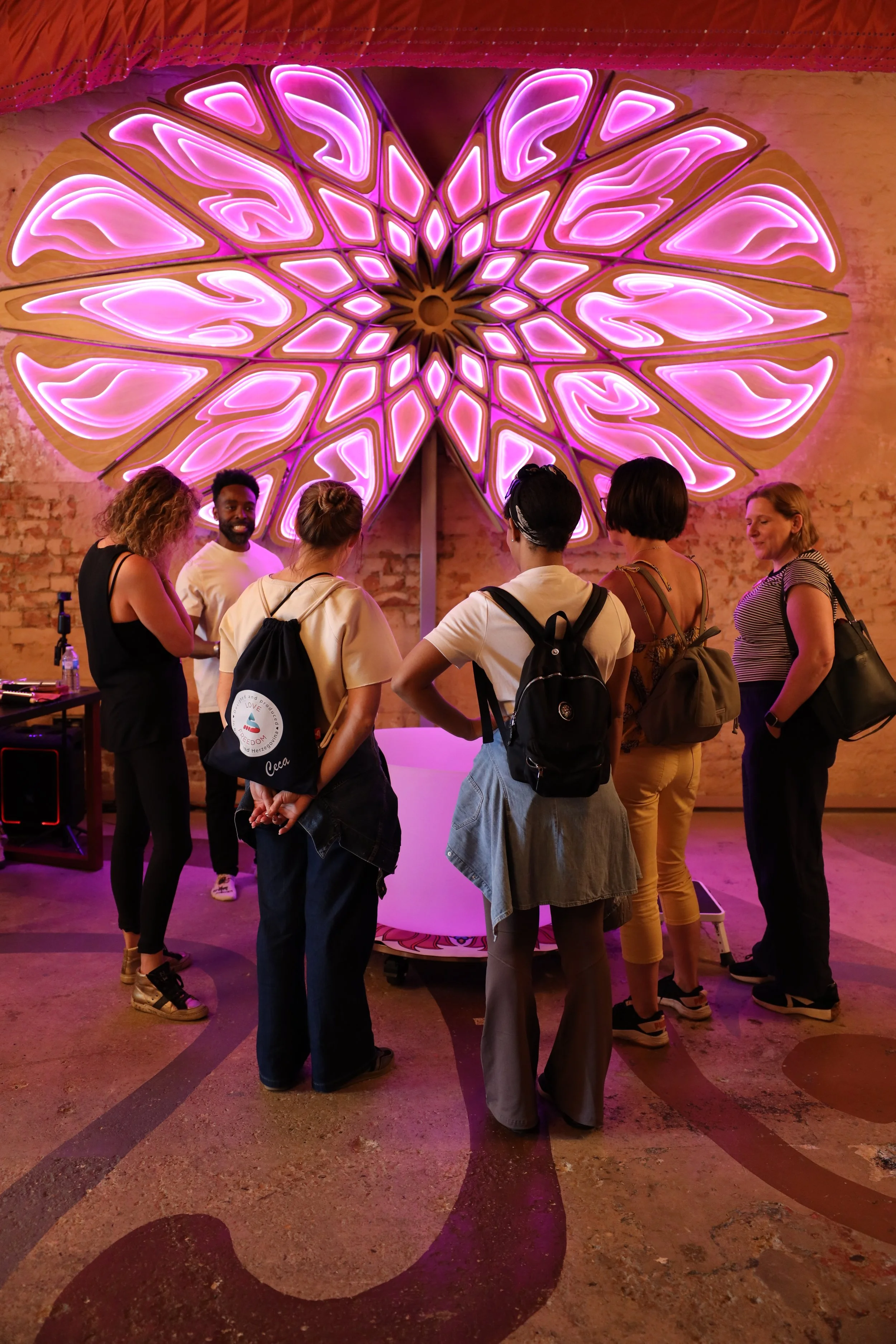 Group of six people gathered around a illuminated pink and purple abstract art piece on a wall in an indoor space with a brick wall background.