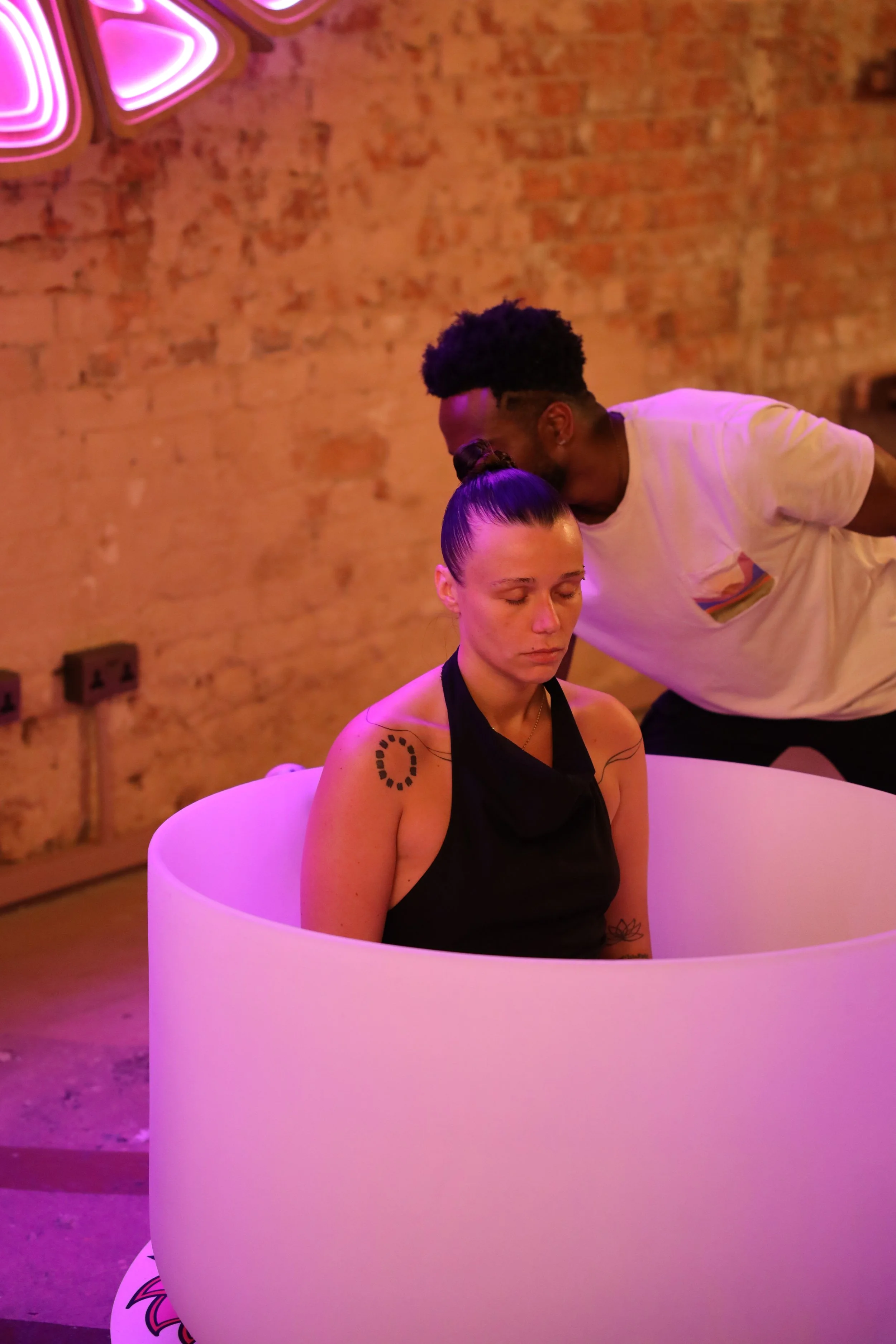 A woman sitting inside a circular white tub with her eyes closed and a man leaning in to kiss her forehead, in a room with an exposed brick wall and neon pink lighting.